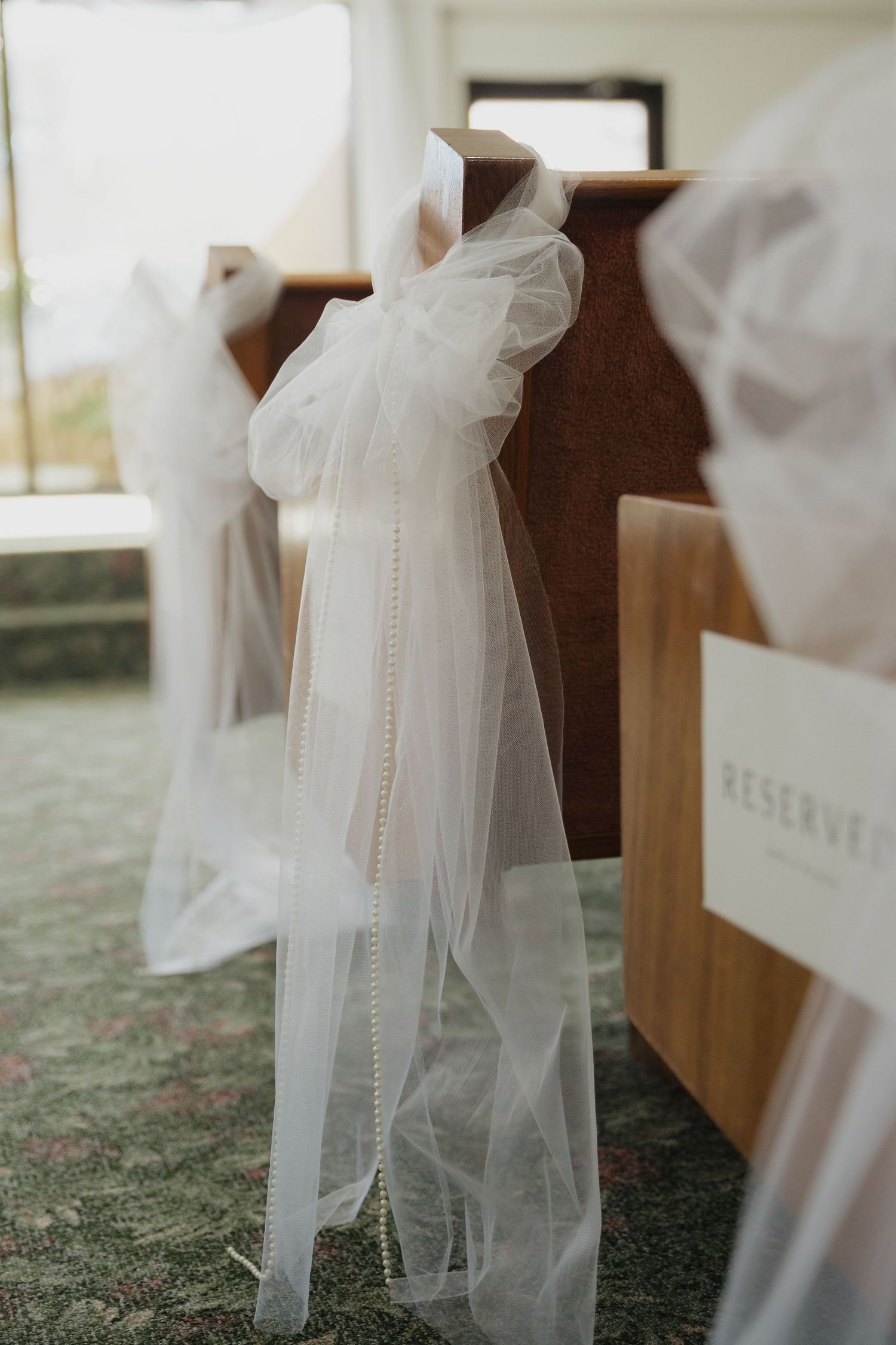 Elegant white tulle and pearl bead bow decorating a wooden church pew at a wedding ceremony.
