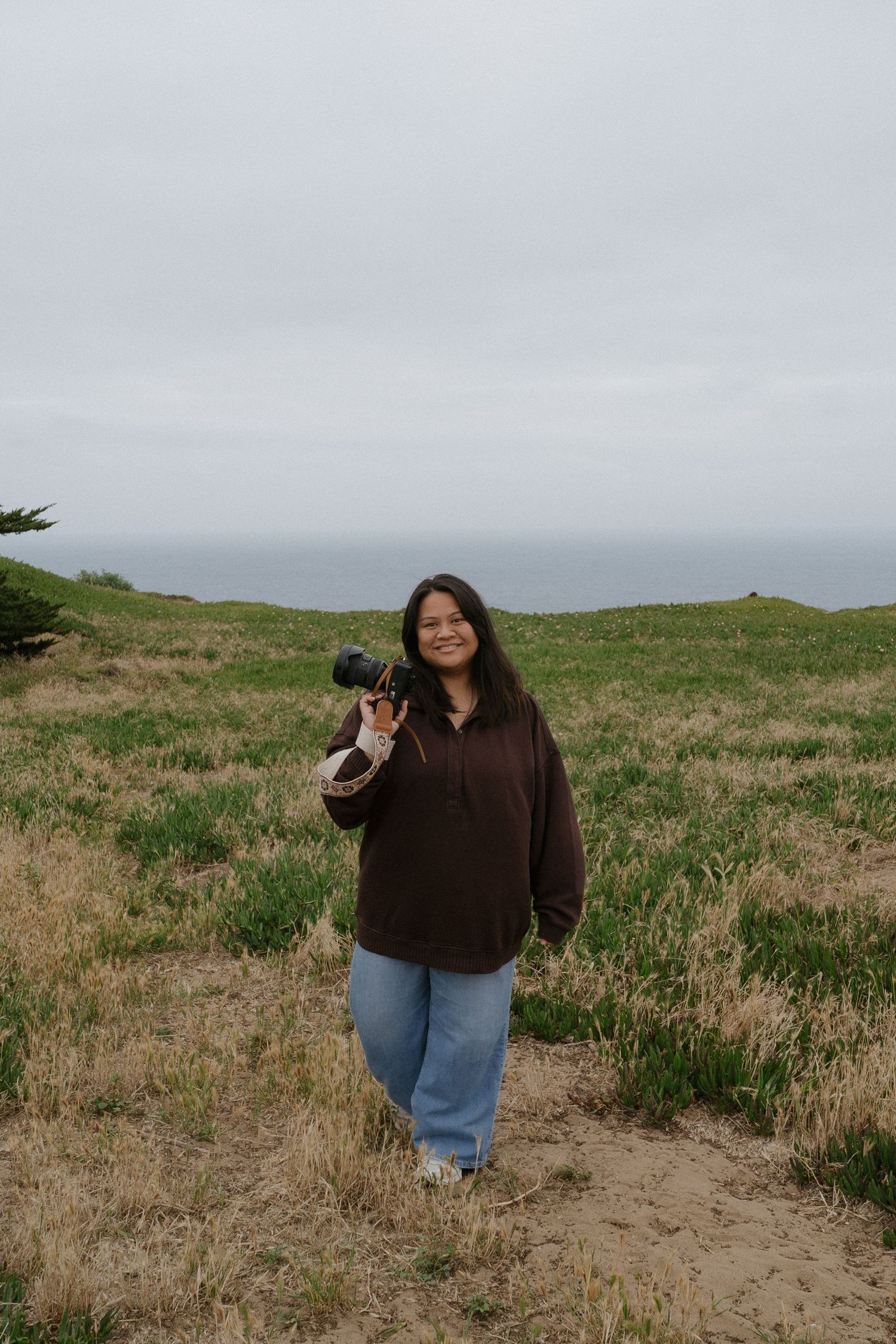 Woman in brown hoodie and jeans holds camera in grassy coastal field under overcast sky.