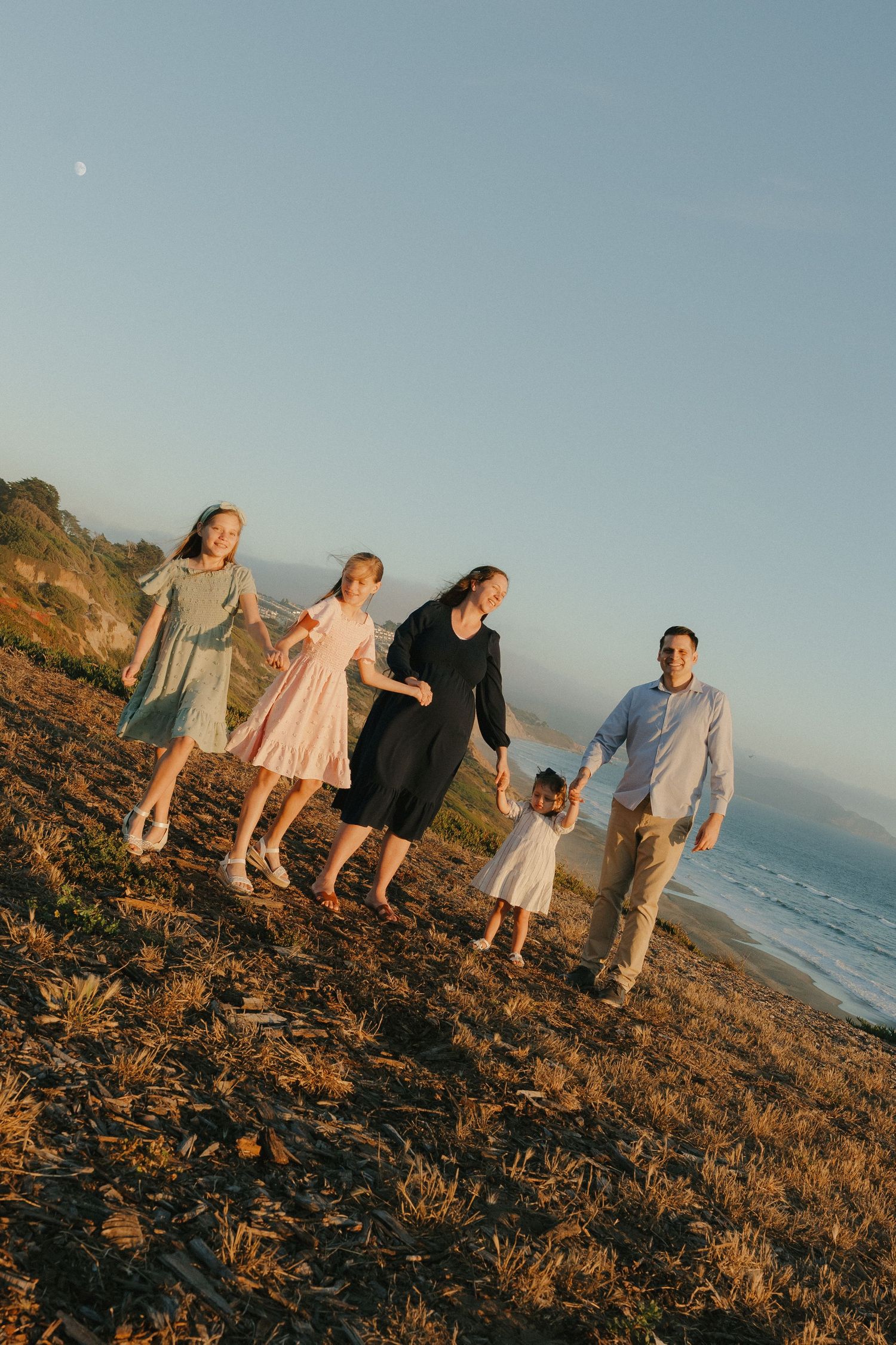 Family of five walking hand-in-hand along a scenic coastal hillside at golden hour sunset.