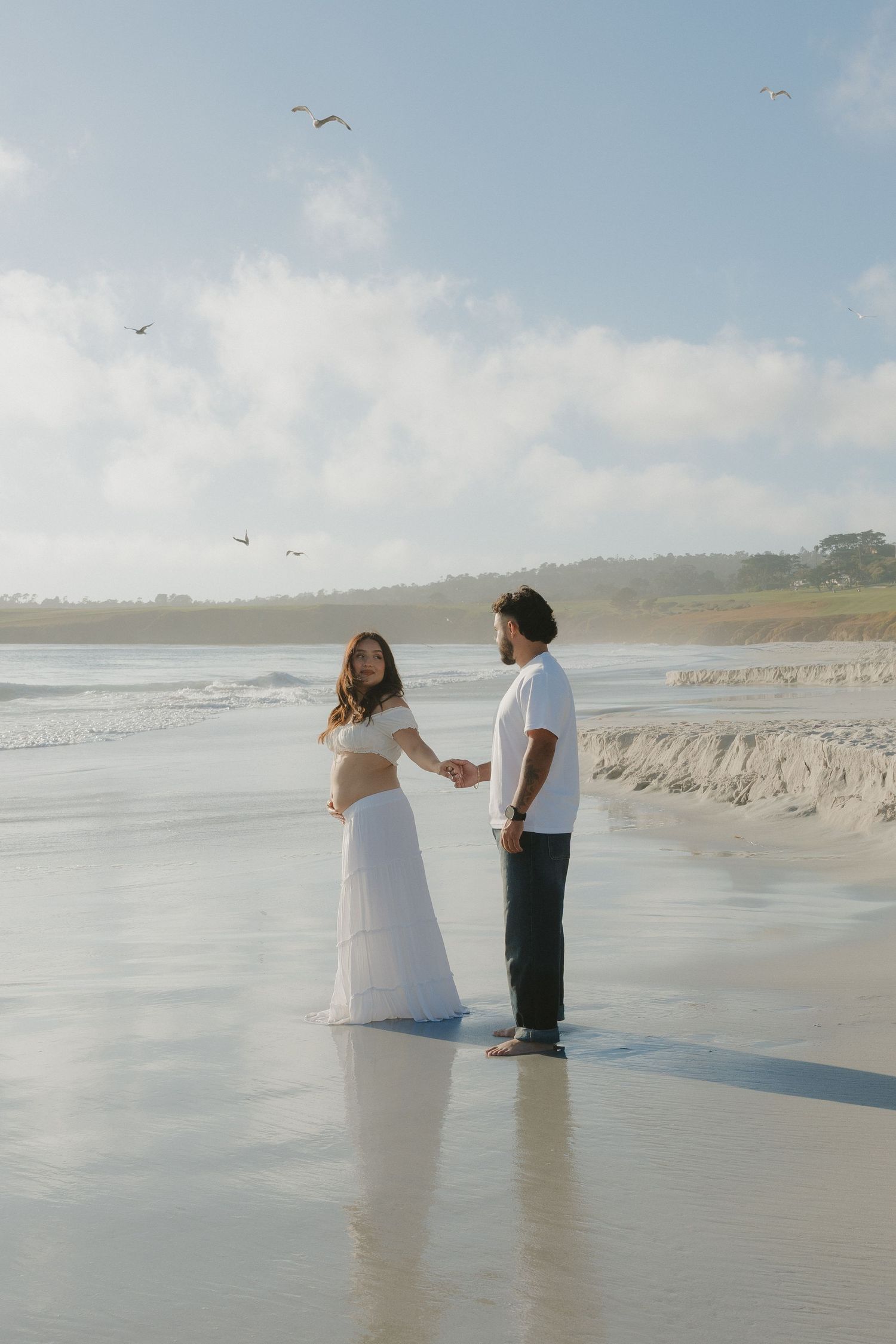 Pregnant couple holding hands on a serene beach, woman in white dress, man in dark pants, with birds flying overhead.