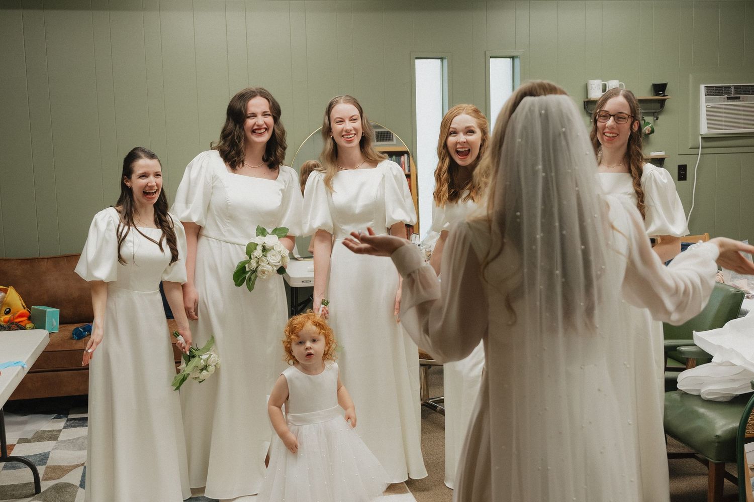 Bridesmaids in white dresses reacting joyfully as bride in veil enters the room with flower girl.