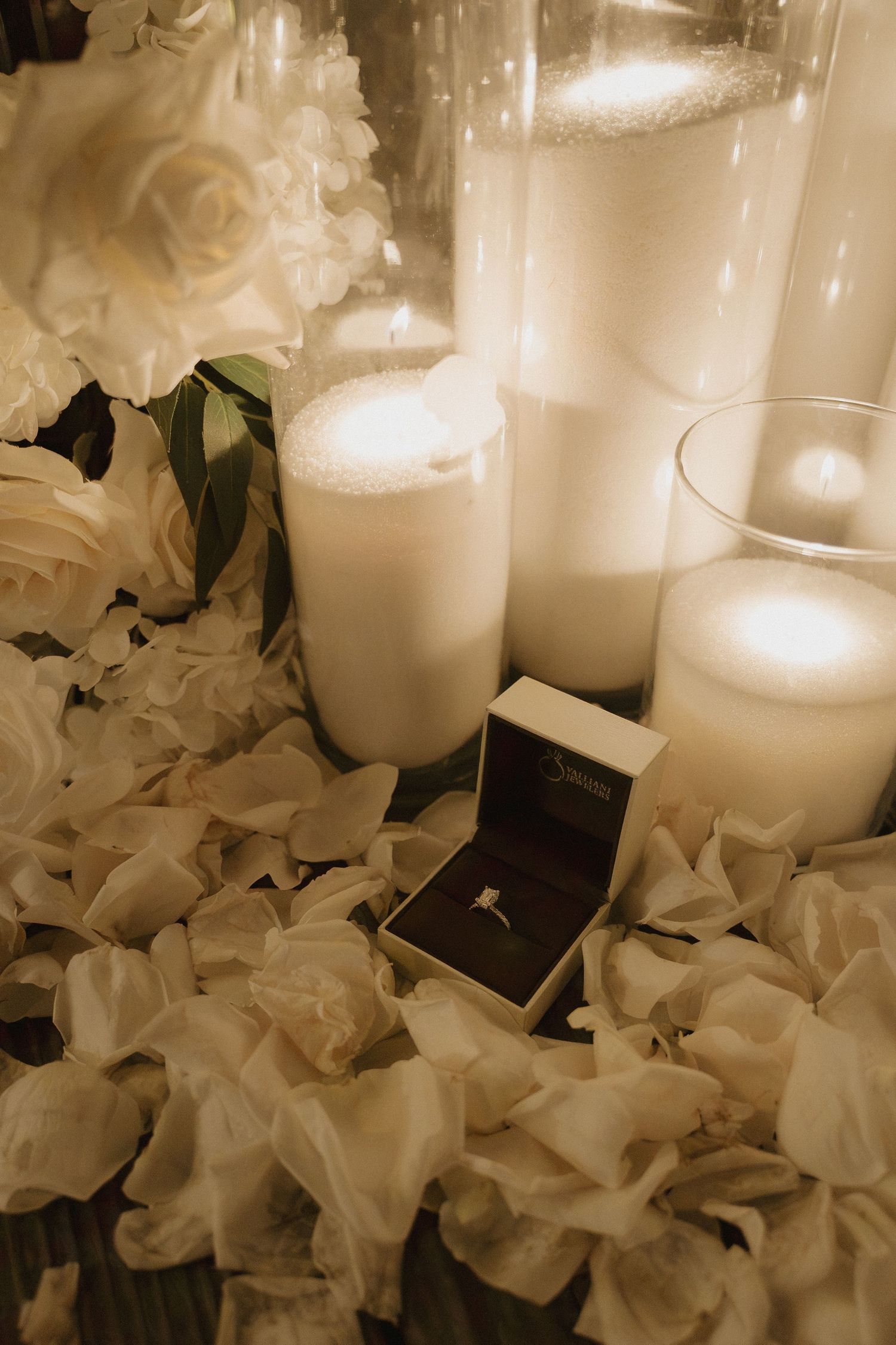 Elegant engagement ring in a box surrounded by white candles and rose petals in a romantic setting.