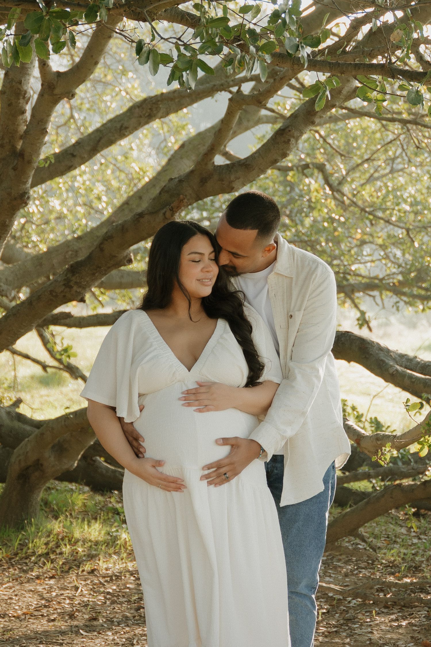 Couple tenderly embracing outdoors, pregnant woman in white dress with partner holding her baby bump beneath sunlit trees in Vacaville, CA