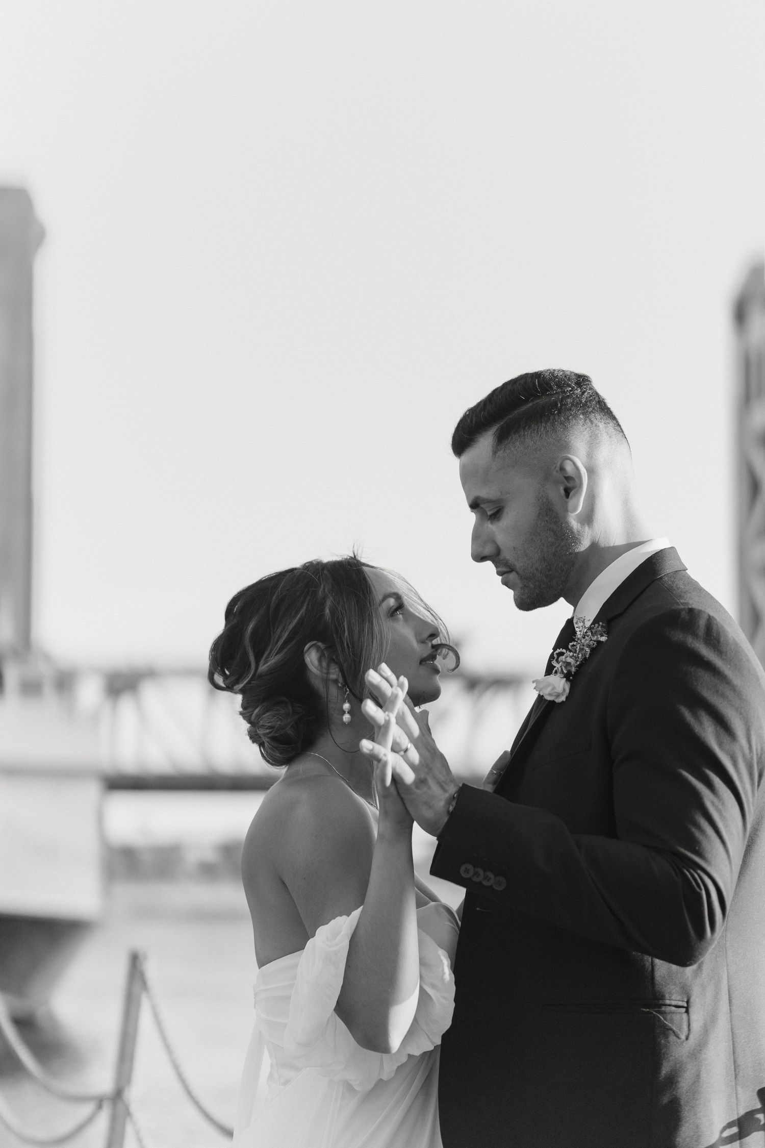 Black and white photo of a bride and groom sharing a tender moment outdoors, with a bridge visible in the background.