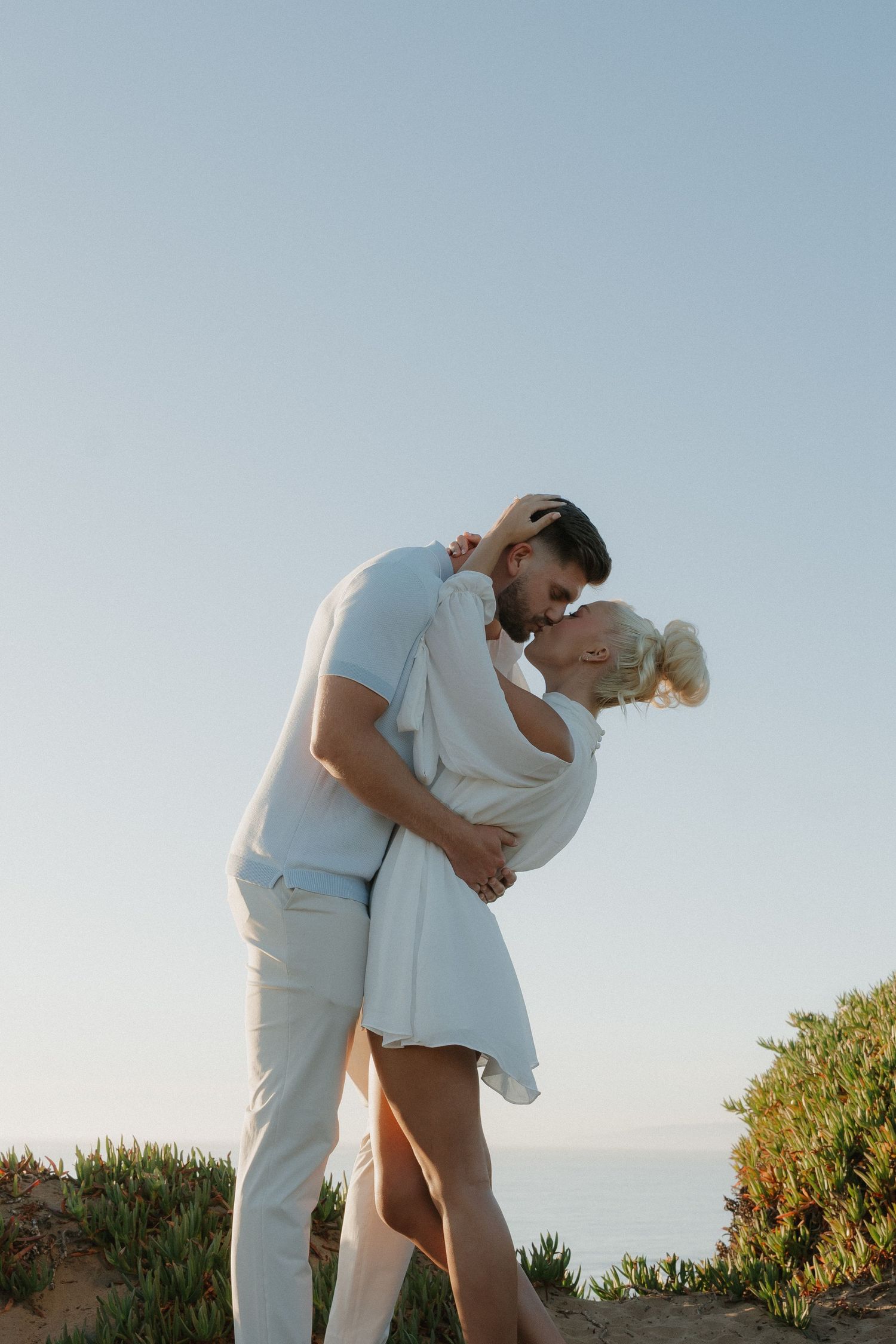 Couple embracing and kissing at golden hour on a coastal cliff overlooking the ocean.
