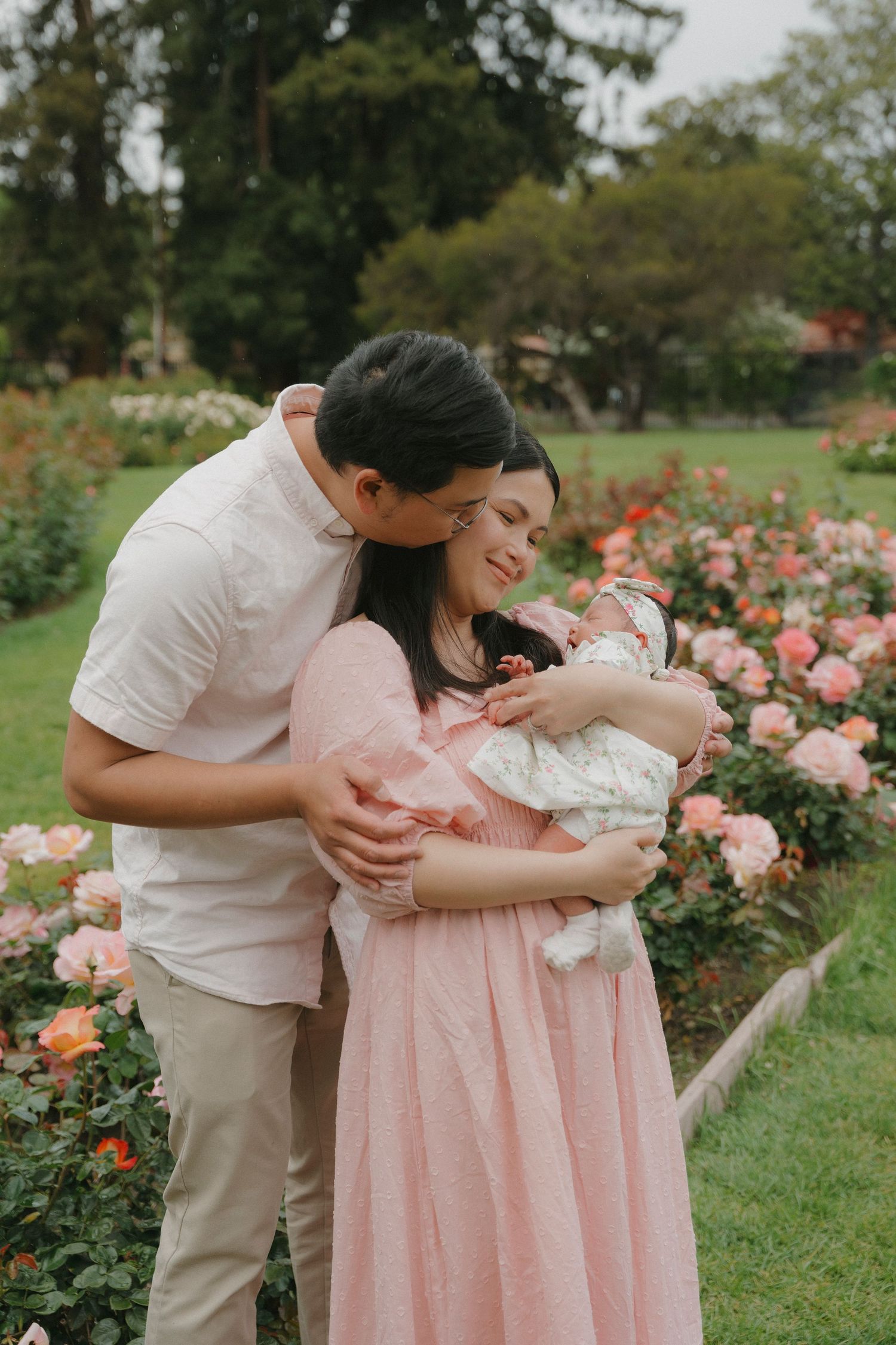 New parents with newborn baby among pink roses in garden, mother in blush dress, father in white shirt at San Jose, CA.