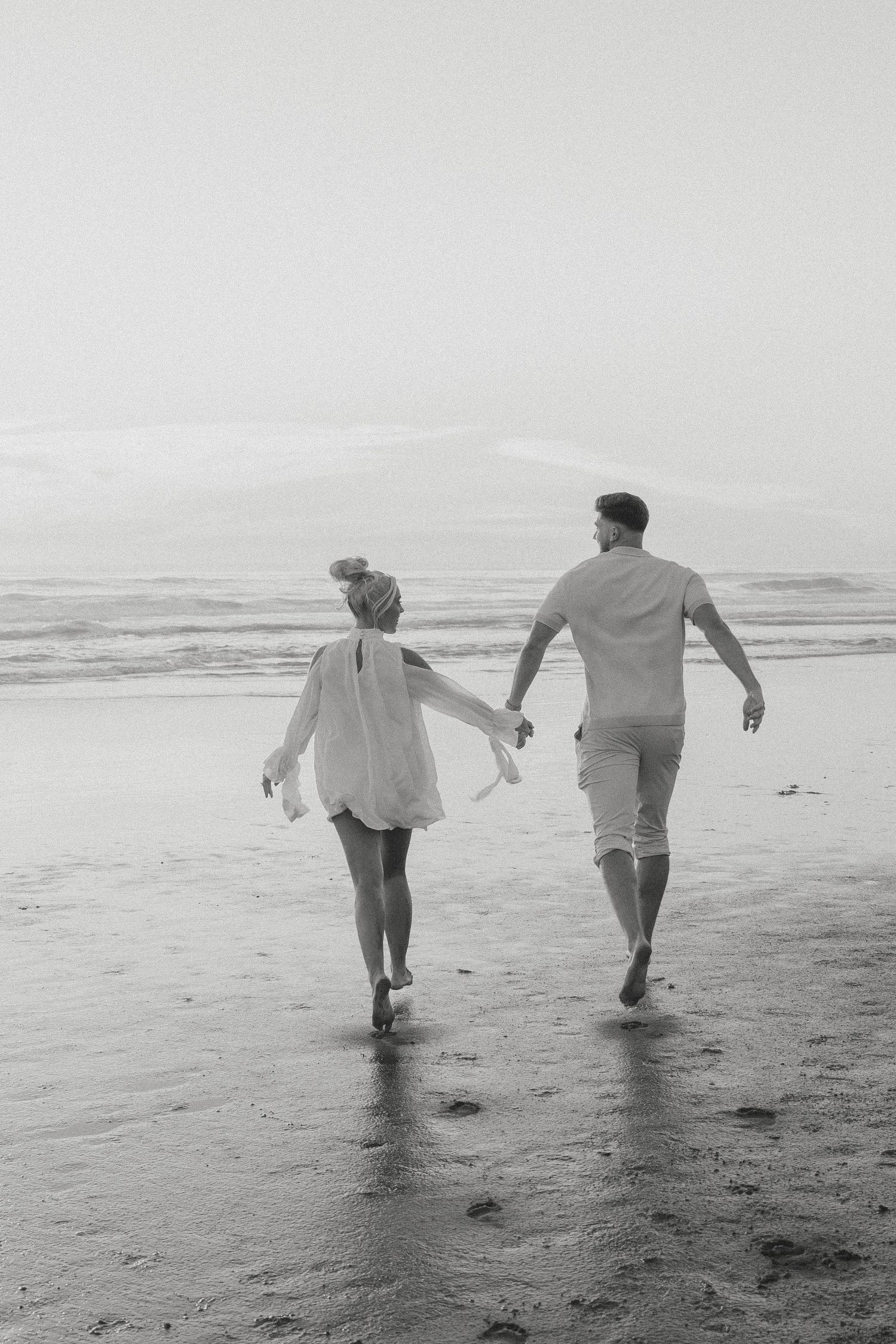 Black and white photo of a couple holding hands while walking along a misty beach shoreline in the Bay Area, CA.