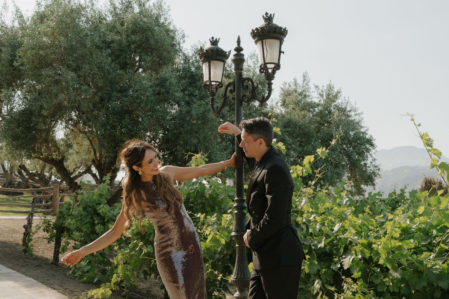 Happy couple dancing together near decorative iron street lamp in a sunny vineyard garden setting.