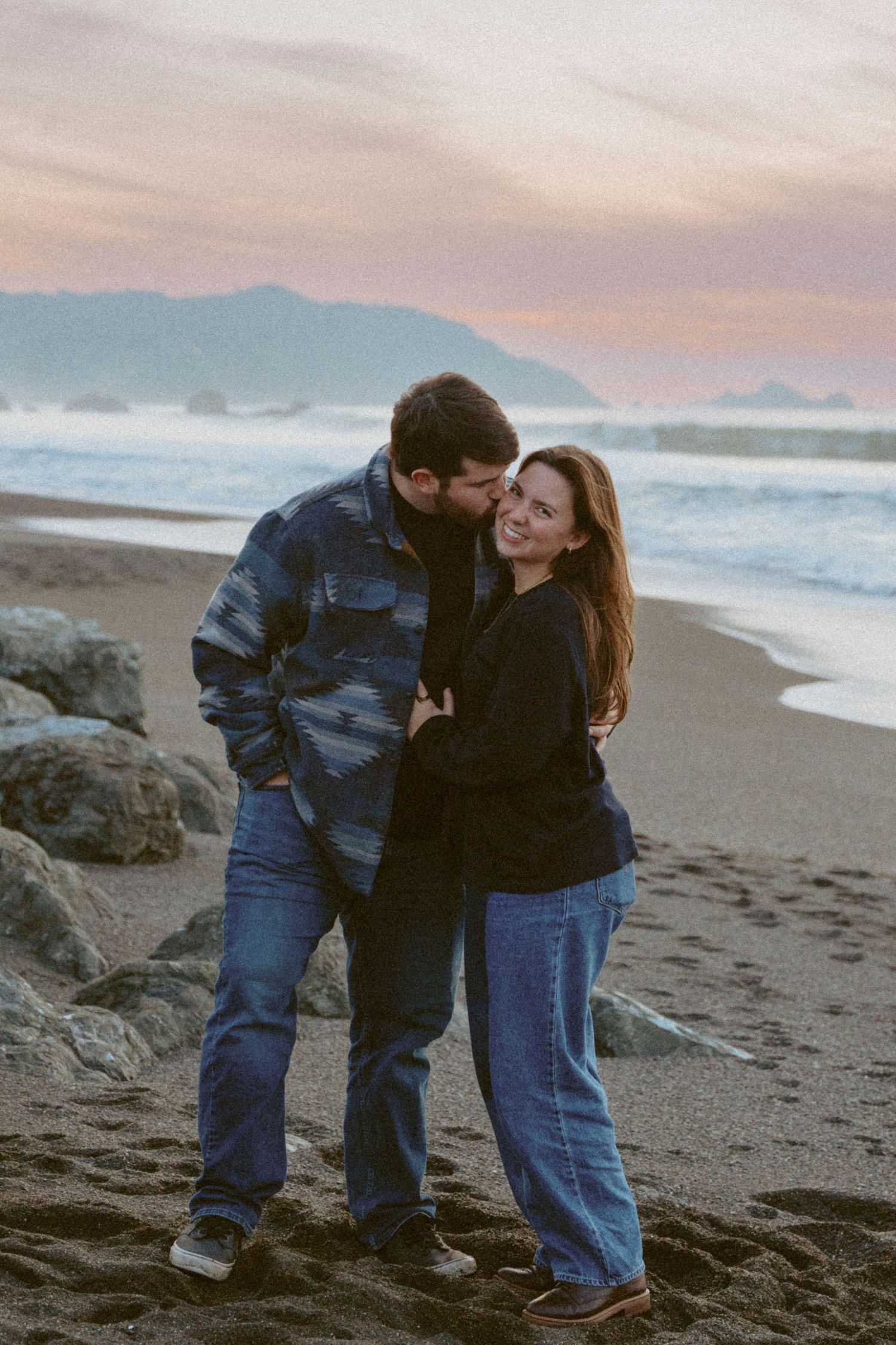 Couple sharing a tender kiss on a dark sandy beach at sunset with ocean waves and mountains behind them at Pacifica, CA.
