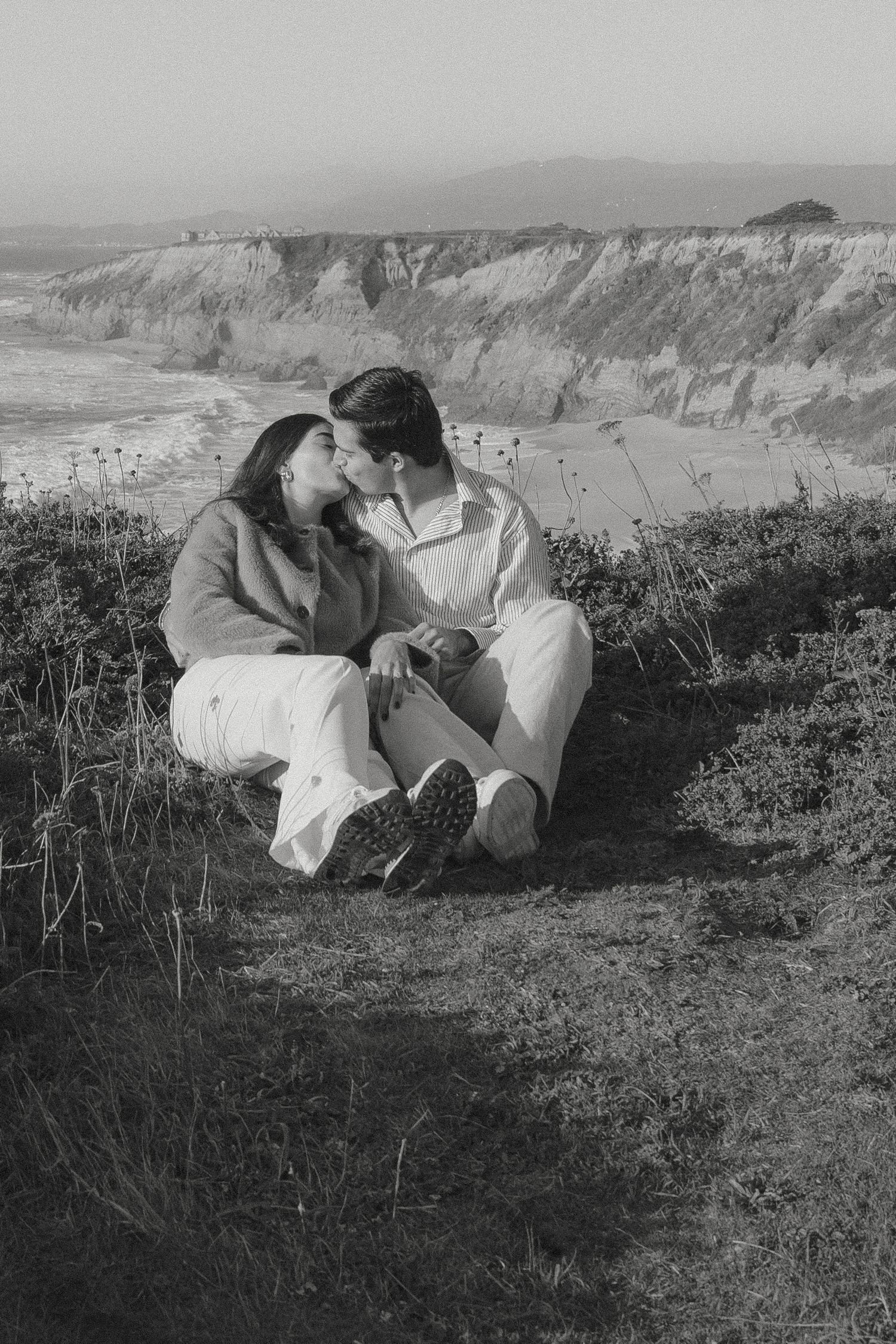 Black and white photo of a couple kissing while sitting on a coastal cliff with ocean and rocky bluffs in background at Half Moon Bay, CA.