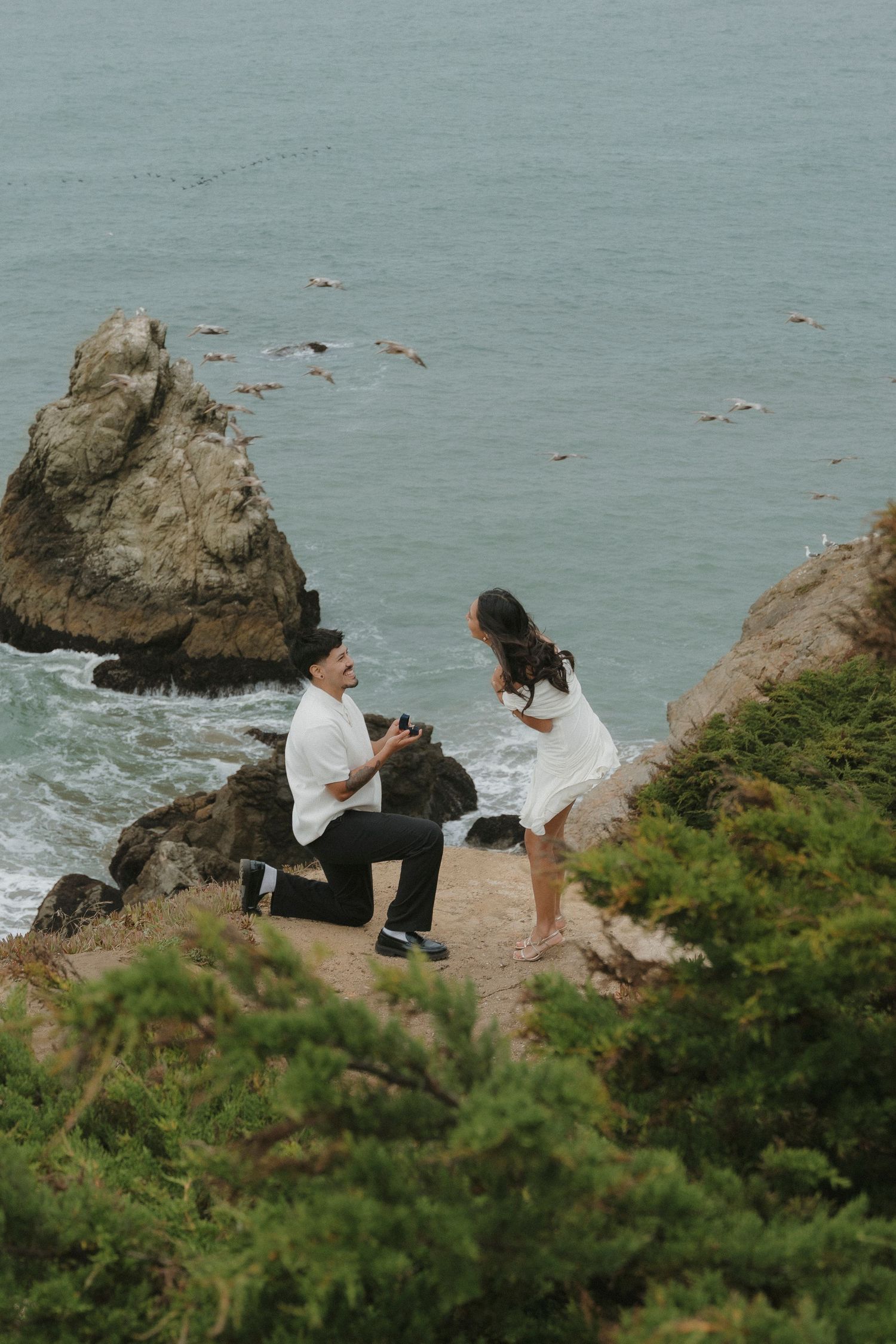Man proposes on one knee to woman on rocky coastal cliff overlooking ocean waves and sea rocks at Lands End.