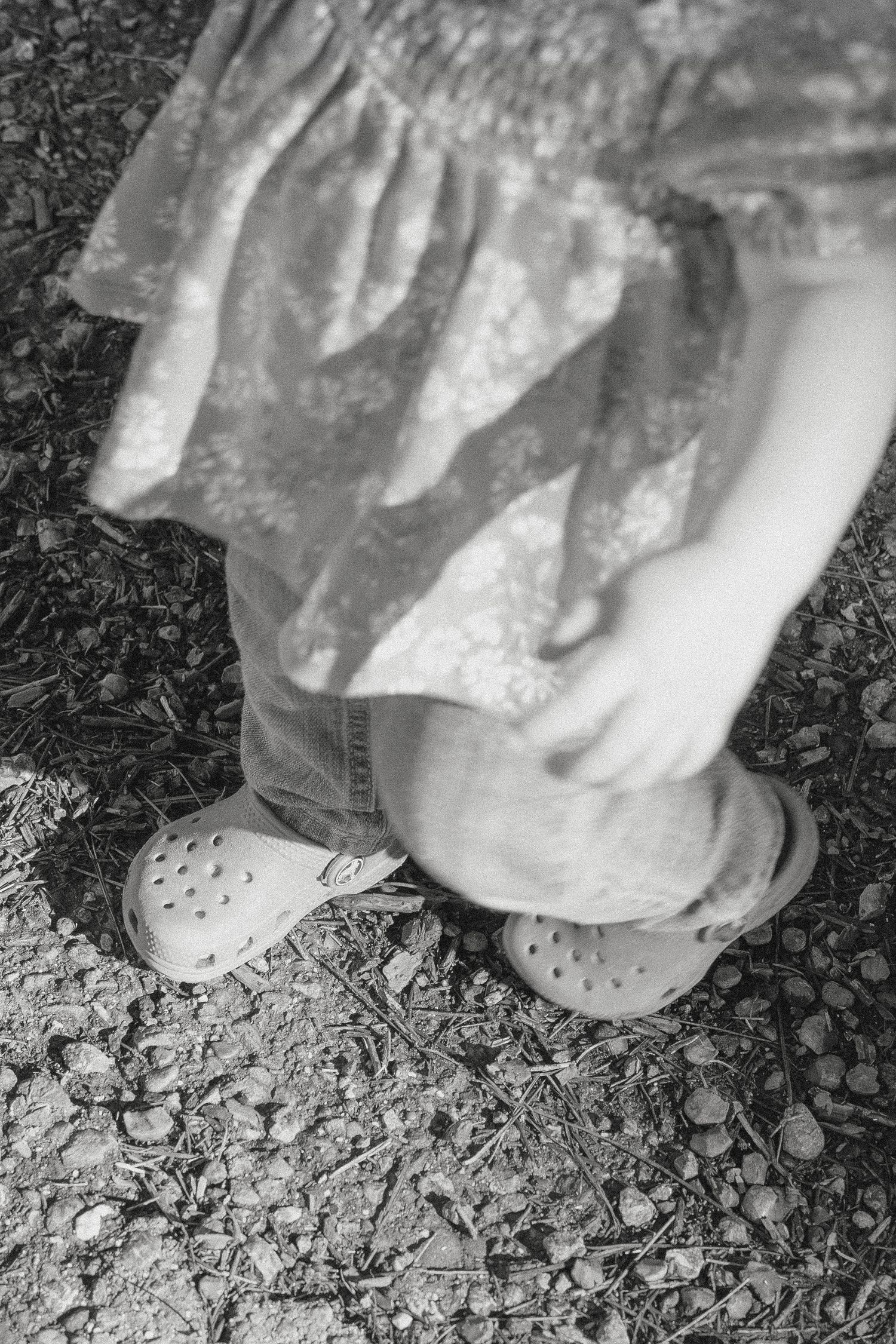 Black and white photo of a child in a floral dress wearing Crocs sandals, standing on a gravel path in Vacaville, CA.