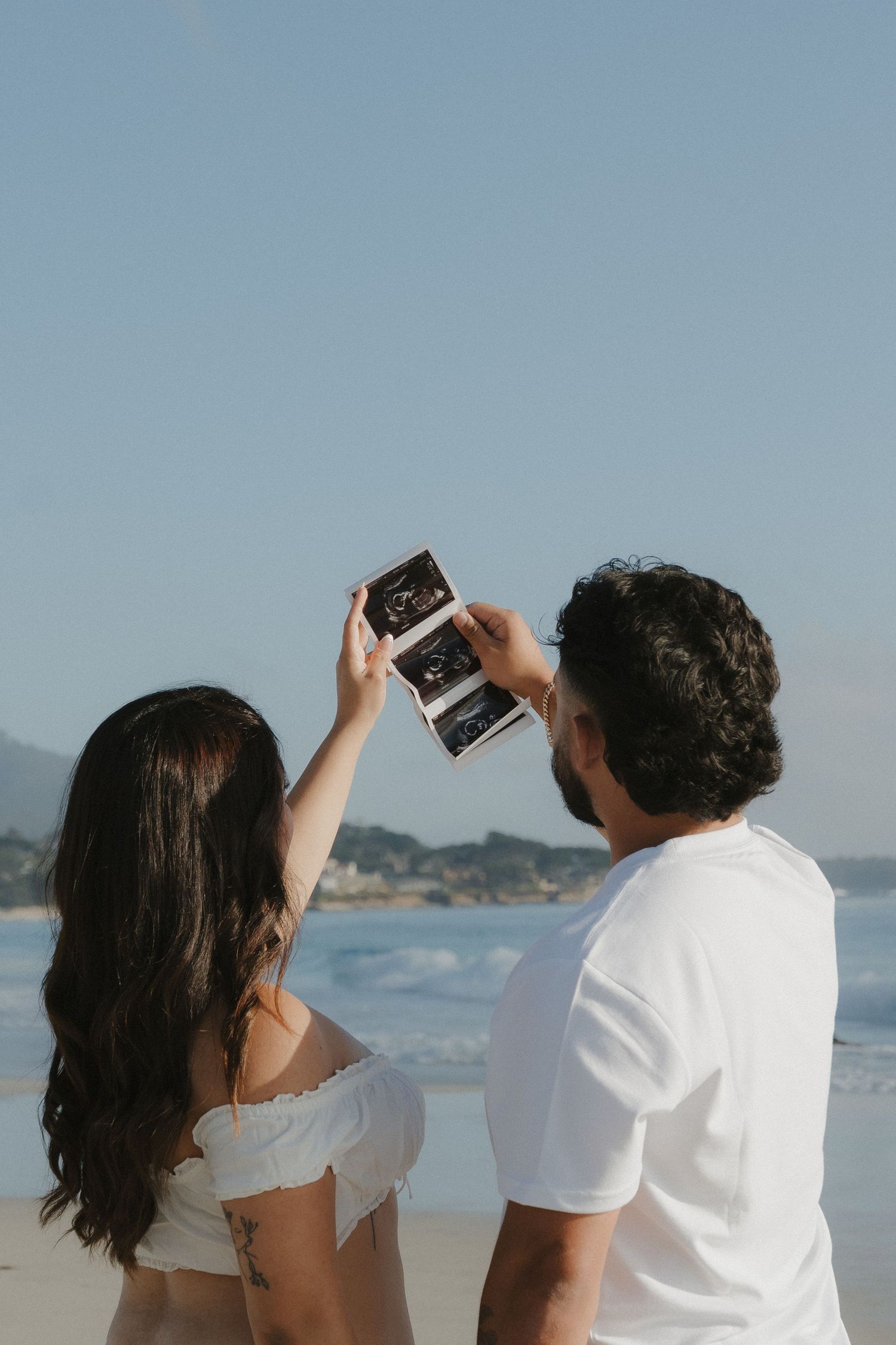 Couple holding ultrasound photos at the beach, sharing a joyful pregnancy announcement moment by the ocean.