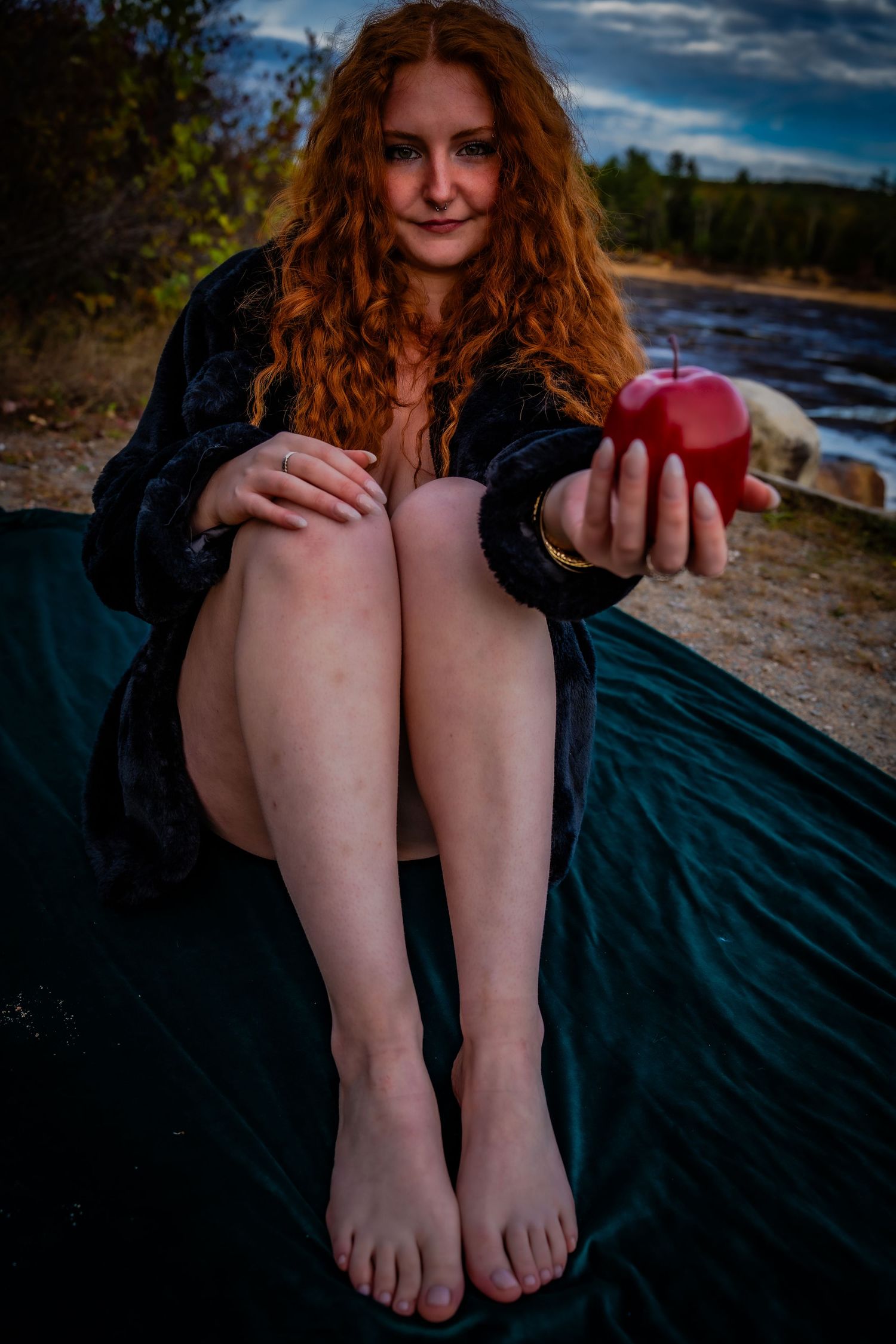 A person with red hair sits on a beach blanket at sunset holding an apple.