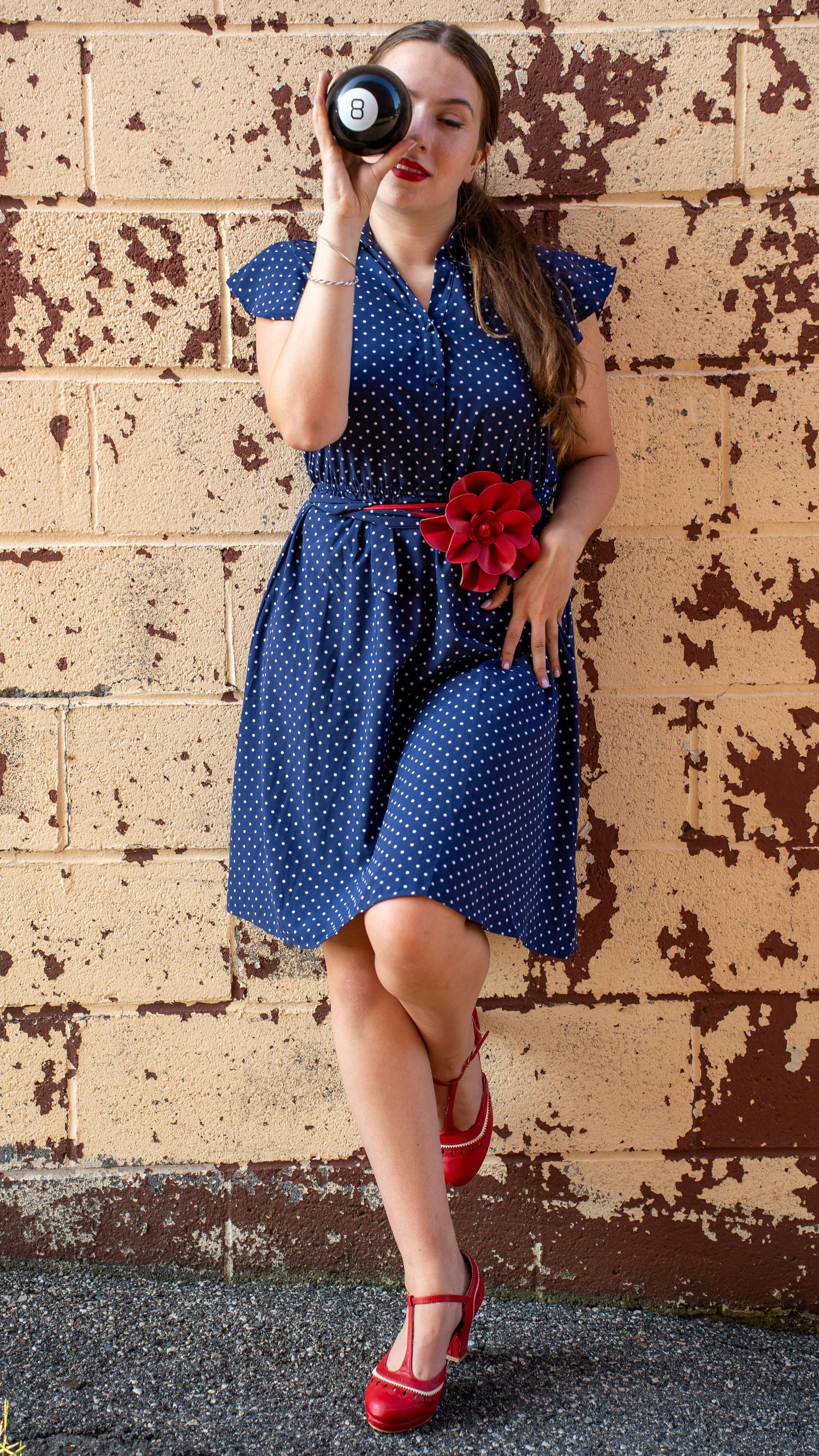 A retro-style polka dot blue dress with red flower accent and matching red heels against a rustic brick wall.
