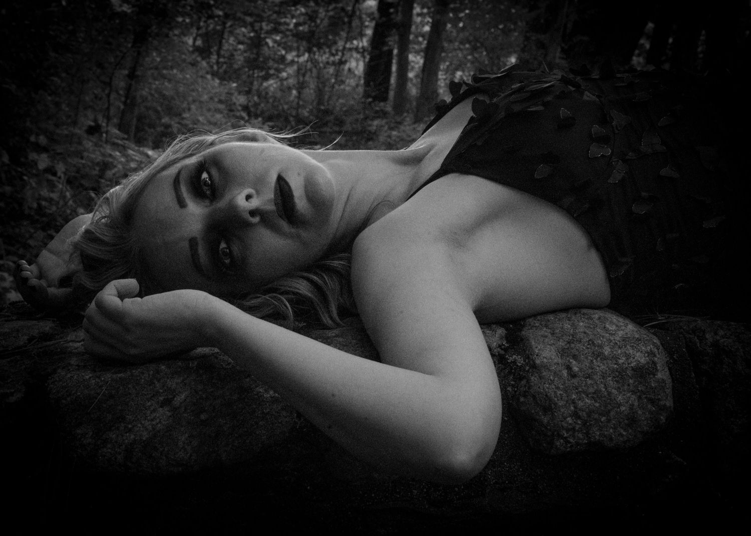 Artistic black and white portrait photograph showing figure reclining on rocks in moody forest setting.