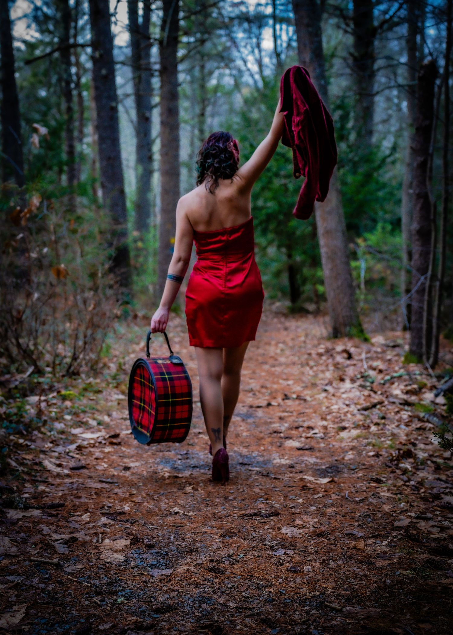 Woman in red dress walking forest path carrying plaid bag and heels in atmospheric evening light.