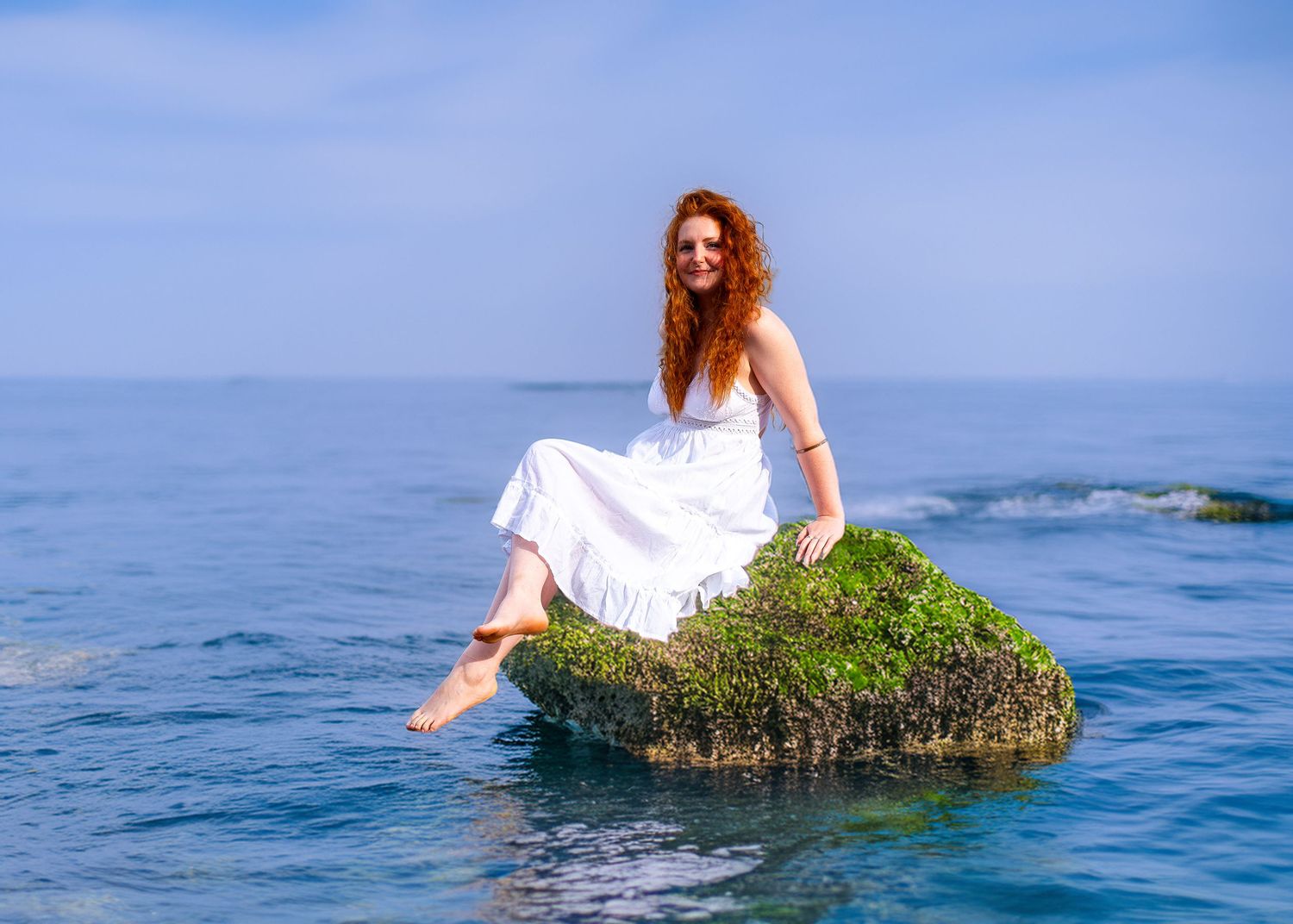 White dress figure perched on mossy rock in blue ocean waters.