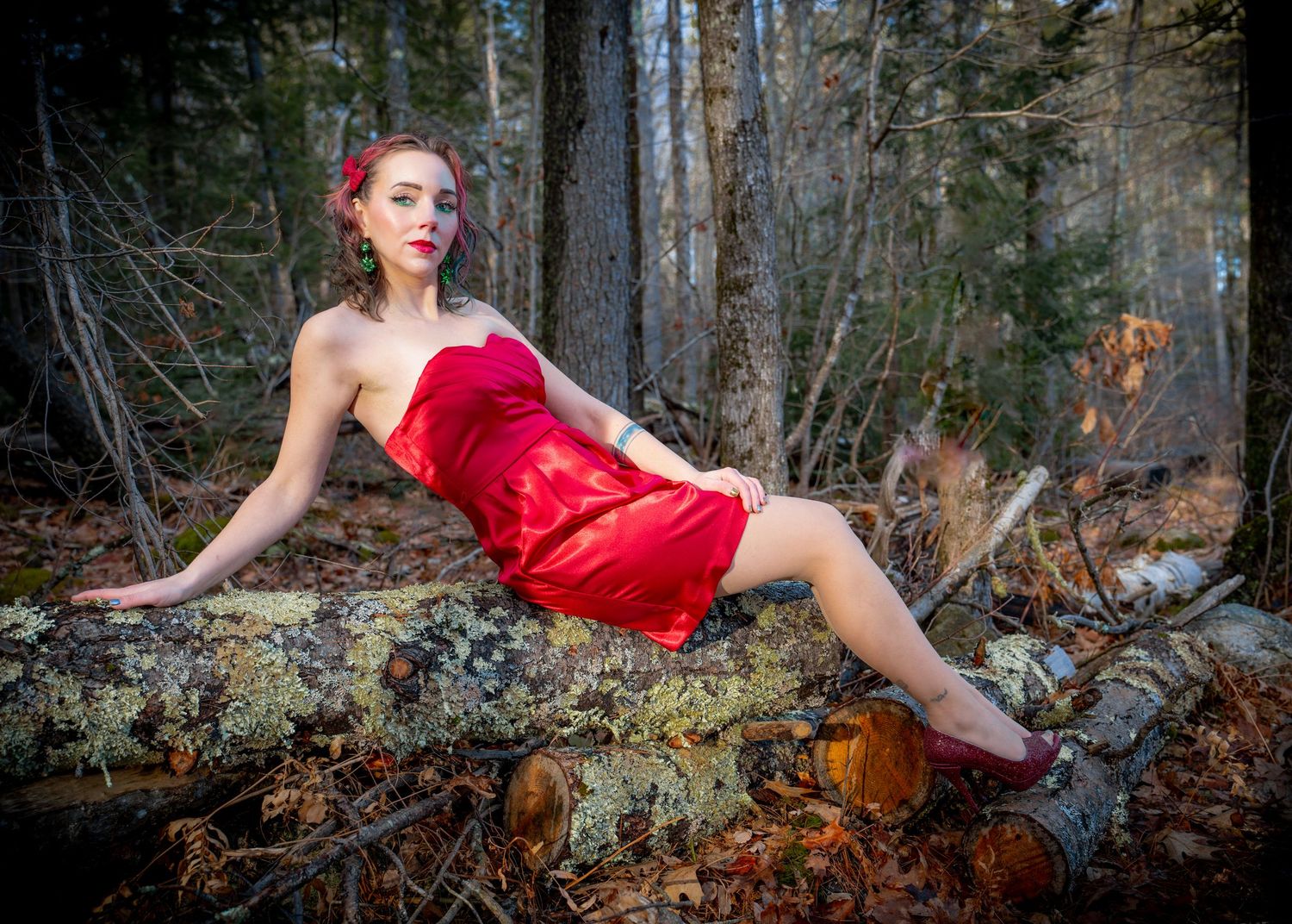A figure in a red dress poses dramatically among fallen leaves in a forest setting.