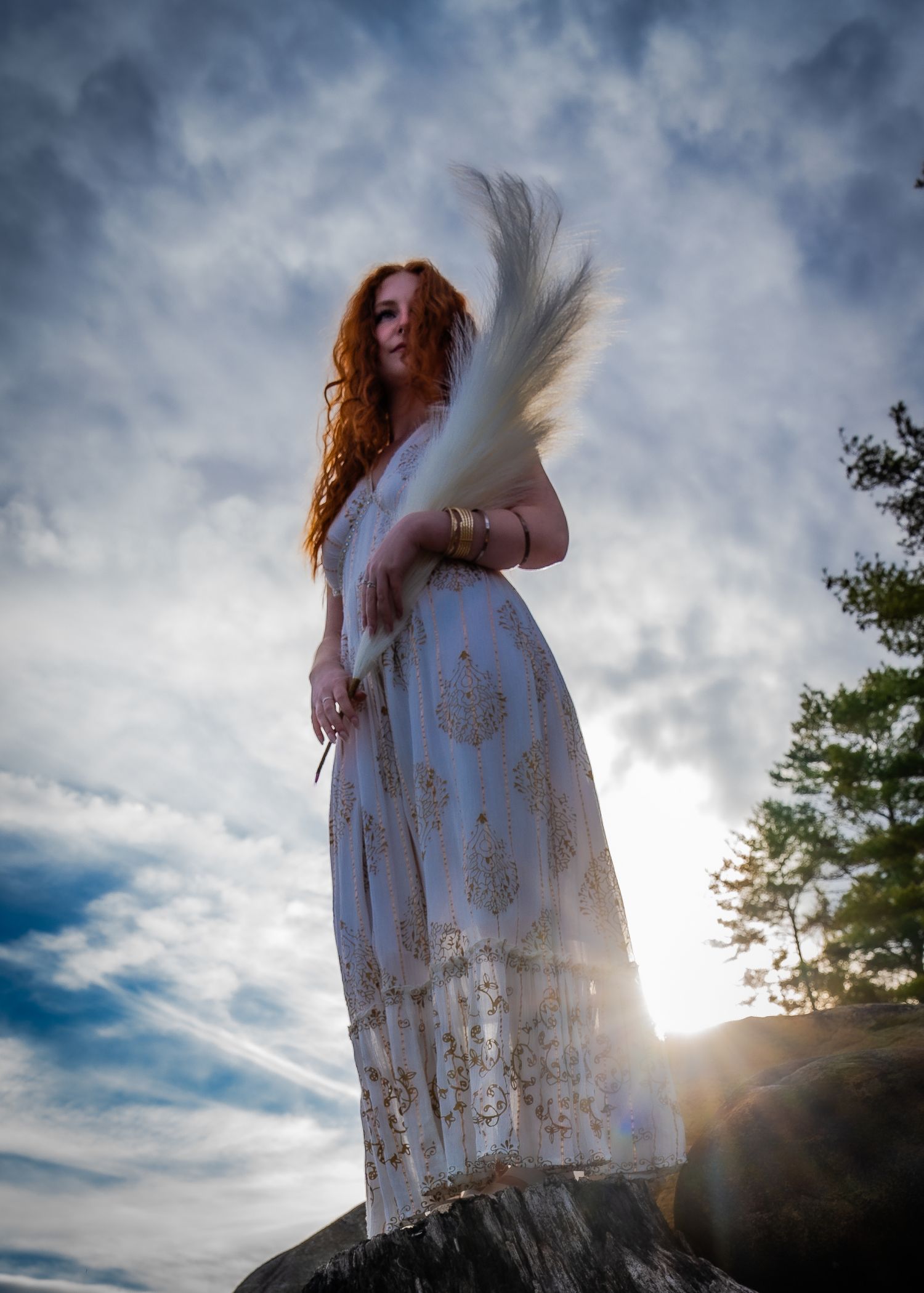 A figure in a white dress stands silhouetted against a dramatic cloudy sky with sunlight breaking through the clouds.