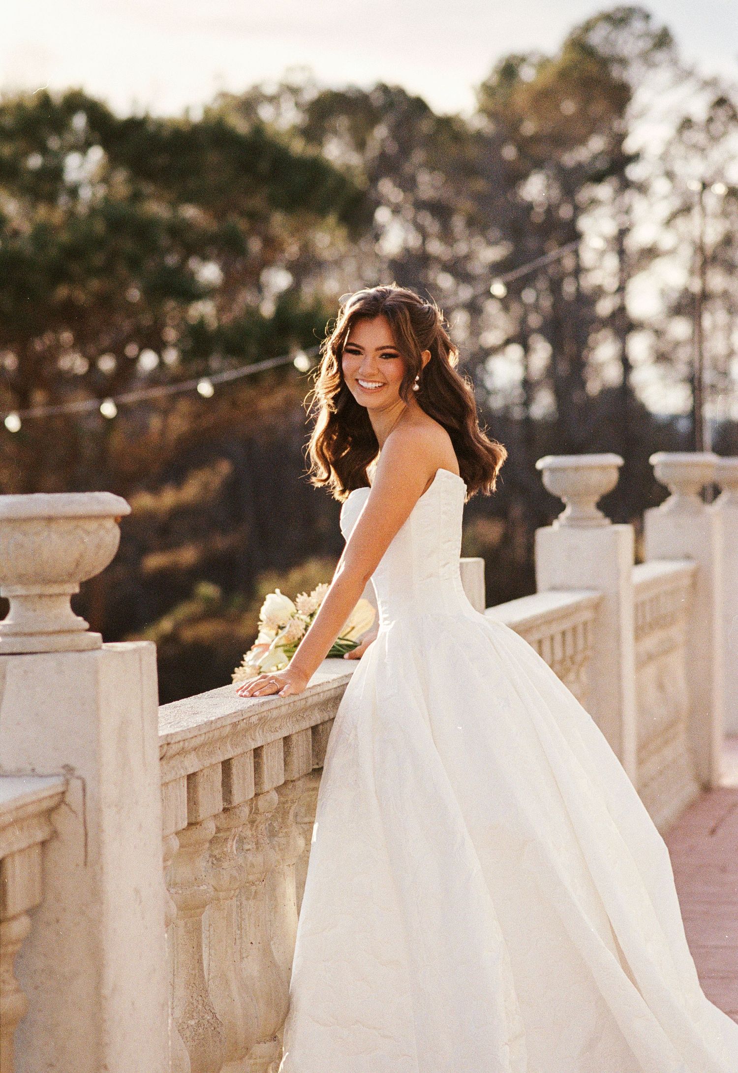Nashville bride smiling on a stone terrace at golden hour, wearing a strapless ballgown and holding a blush bouquet.