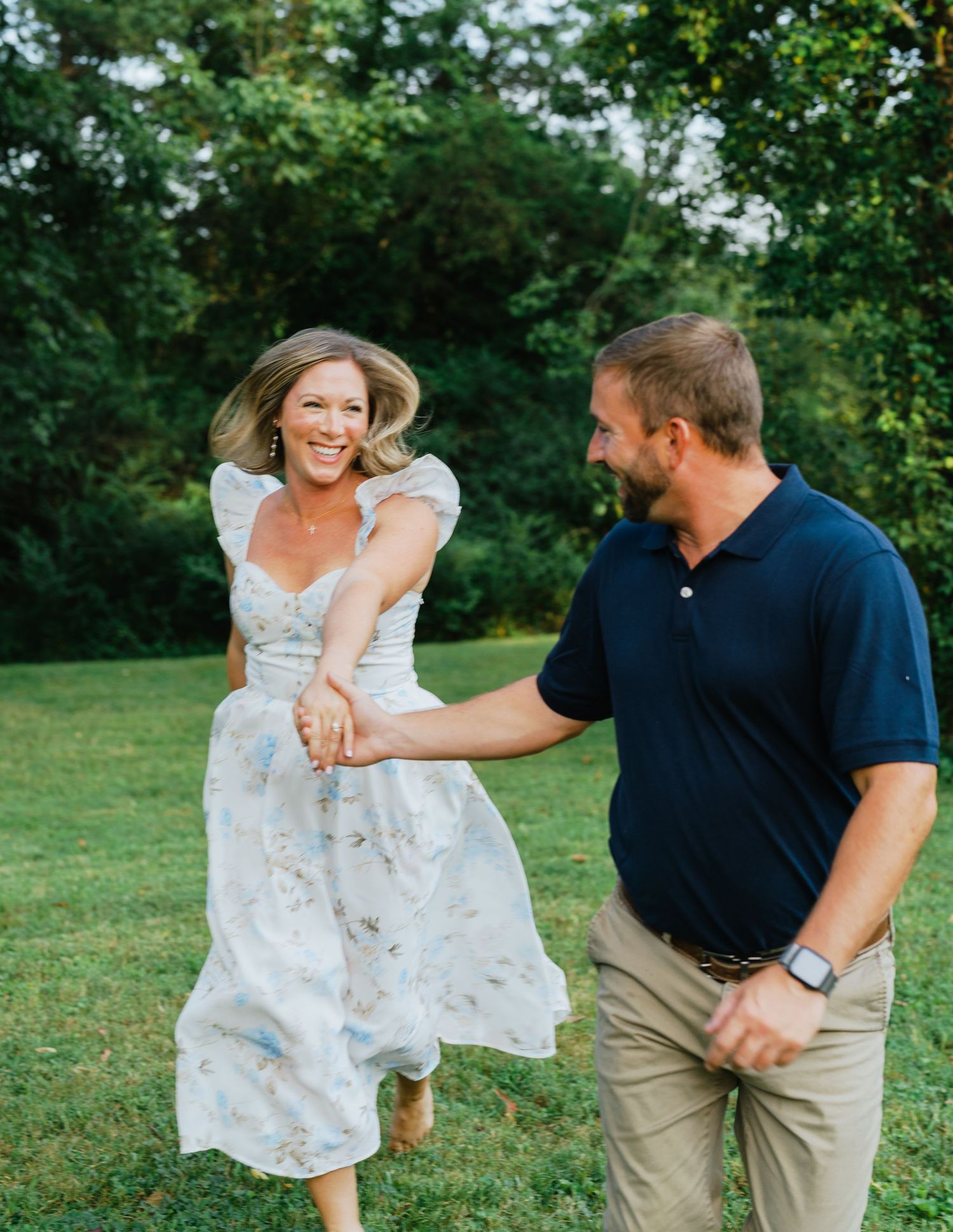 Happy woman in floral dress twirls joyfully with man in green park during engagement session at The Macca Villa at Riverside in Murfreesboro