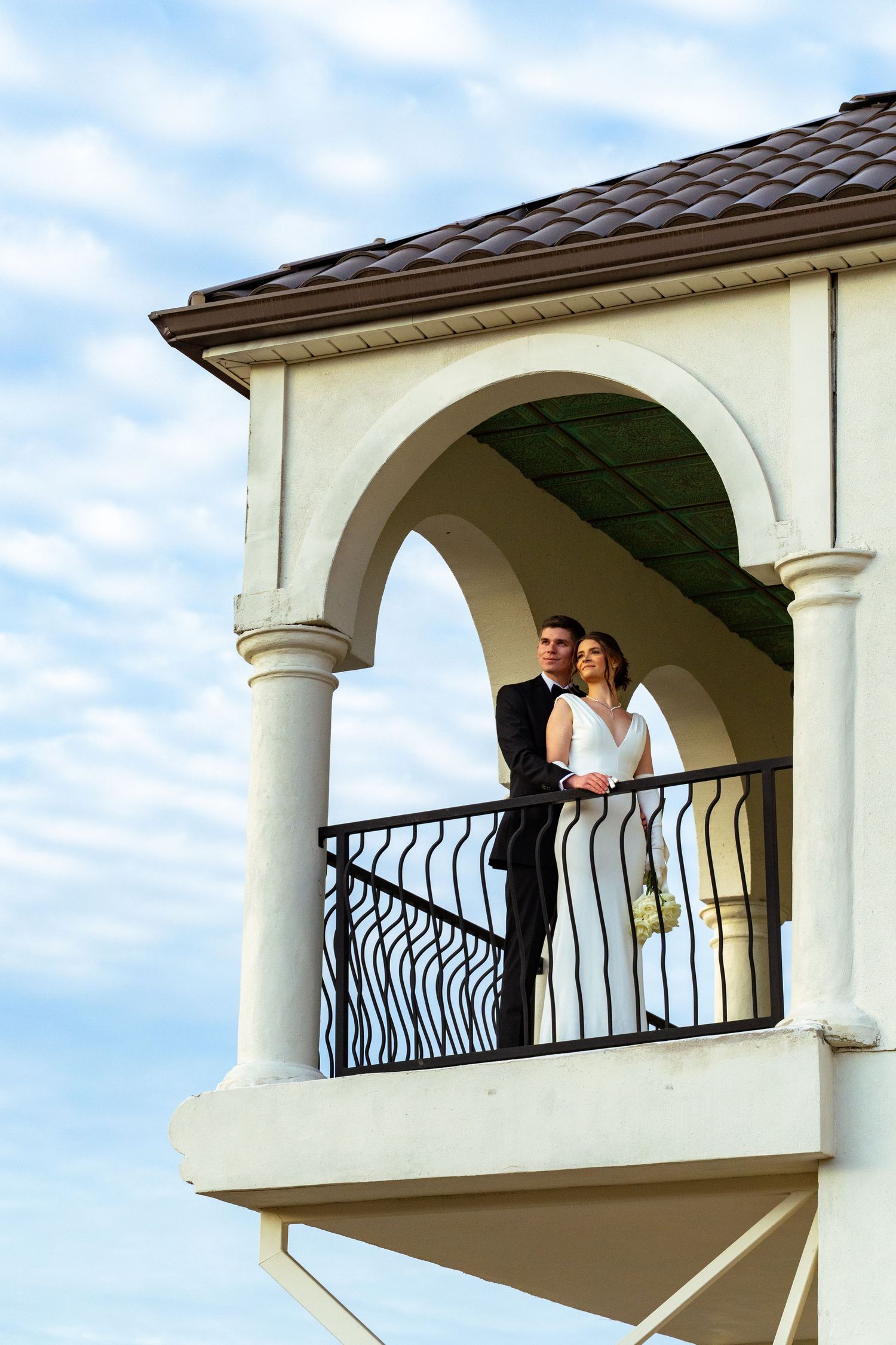 Cookeville wedding couple embracing on a balcony beneath arched columns at sunset, bride in a sleek gown and groom in a black suit.