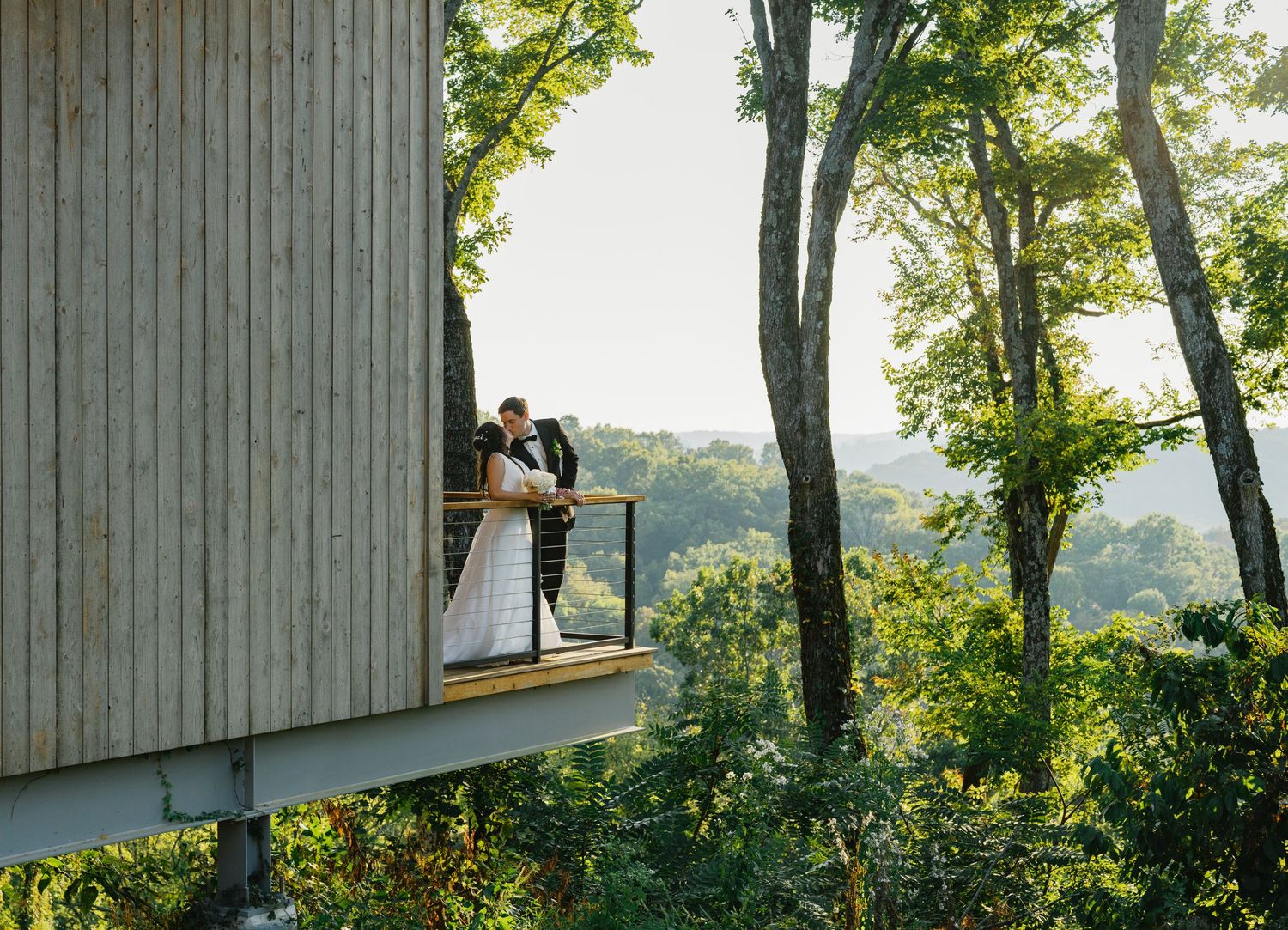 Newlyweds share a romantic kiss on a cantilevered balcony surrounded by lush forest trees at South Hall in Franklin Tennessee.