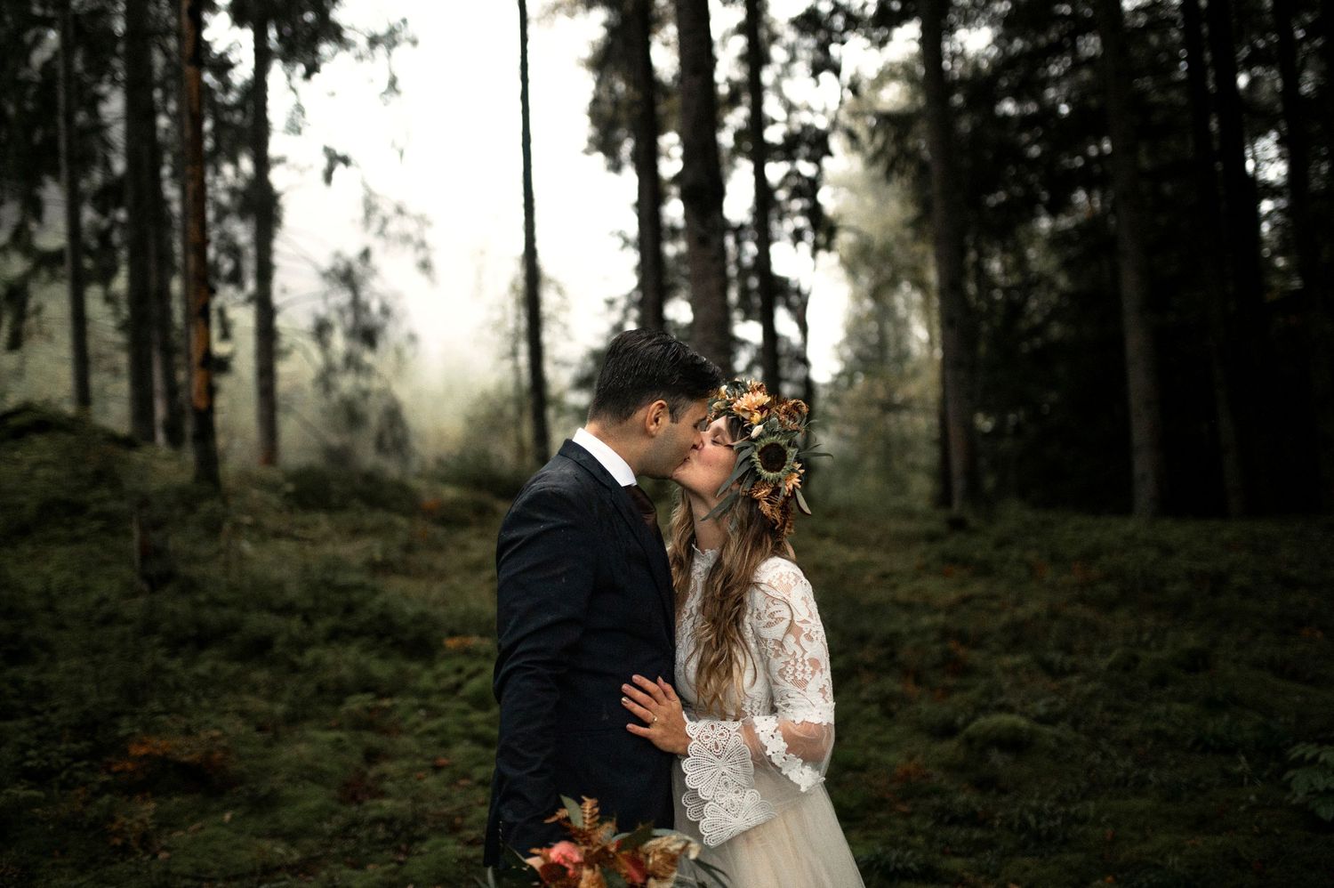 Bride and groom sharing a romantic kiss in a misty forest, bride wearing floral crown and lace dress.