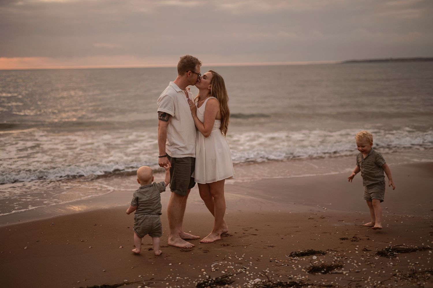 Family of four enjoying a romantic beach sunset, two toddlers playing in sand as parents share a kiss.