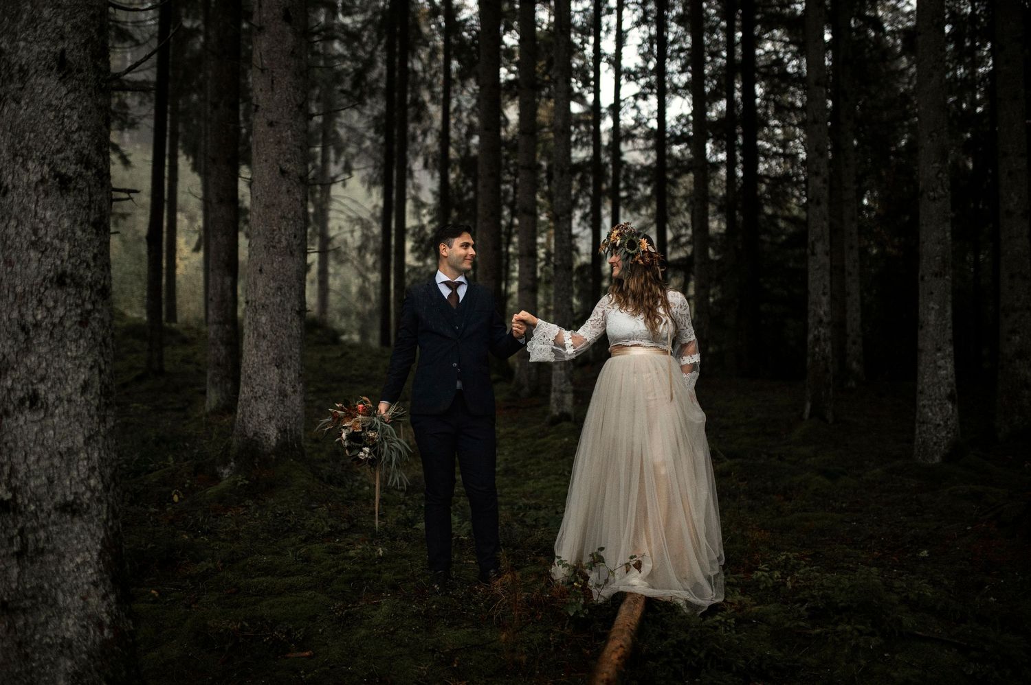 Couple holding hands in a misty forest wedding, bride in white gown and groom in navy suit among tall pine trees. Elopement in Skåne, Sweden