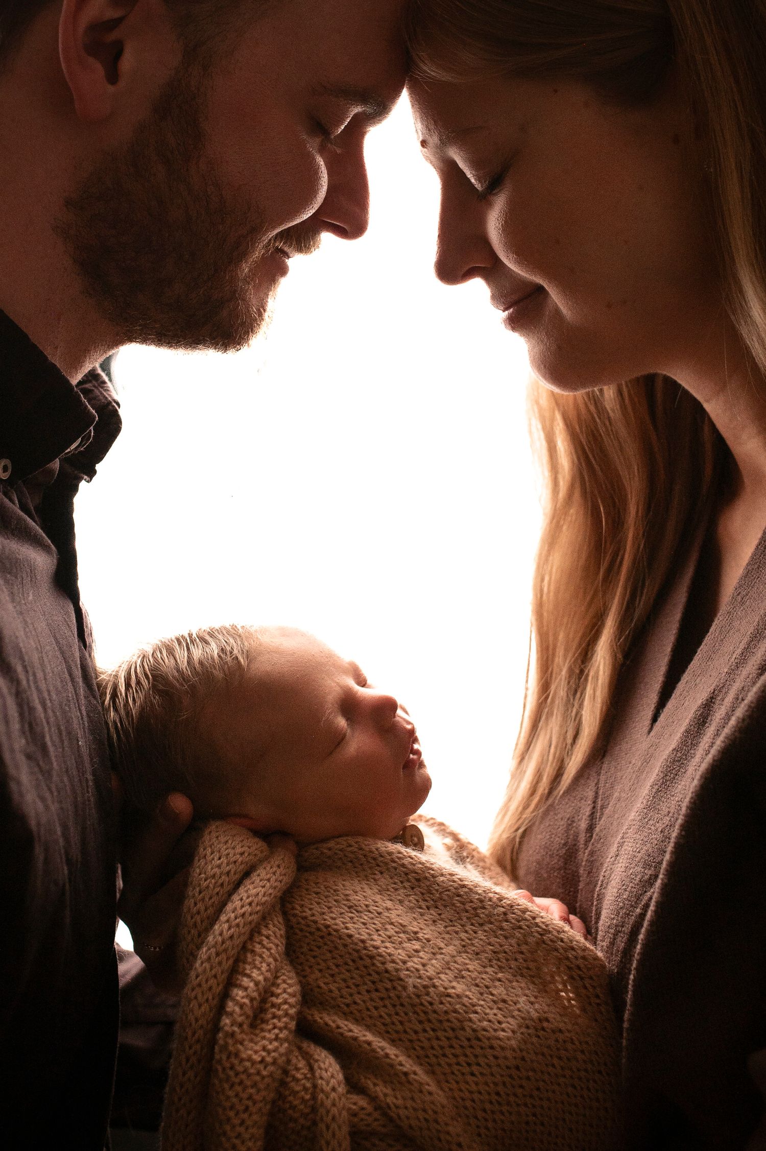 Silhouetted parents tenderly touch foreheads while cradling their sleeping newborn in warm light.