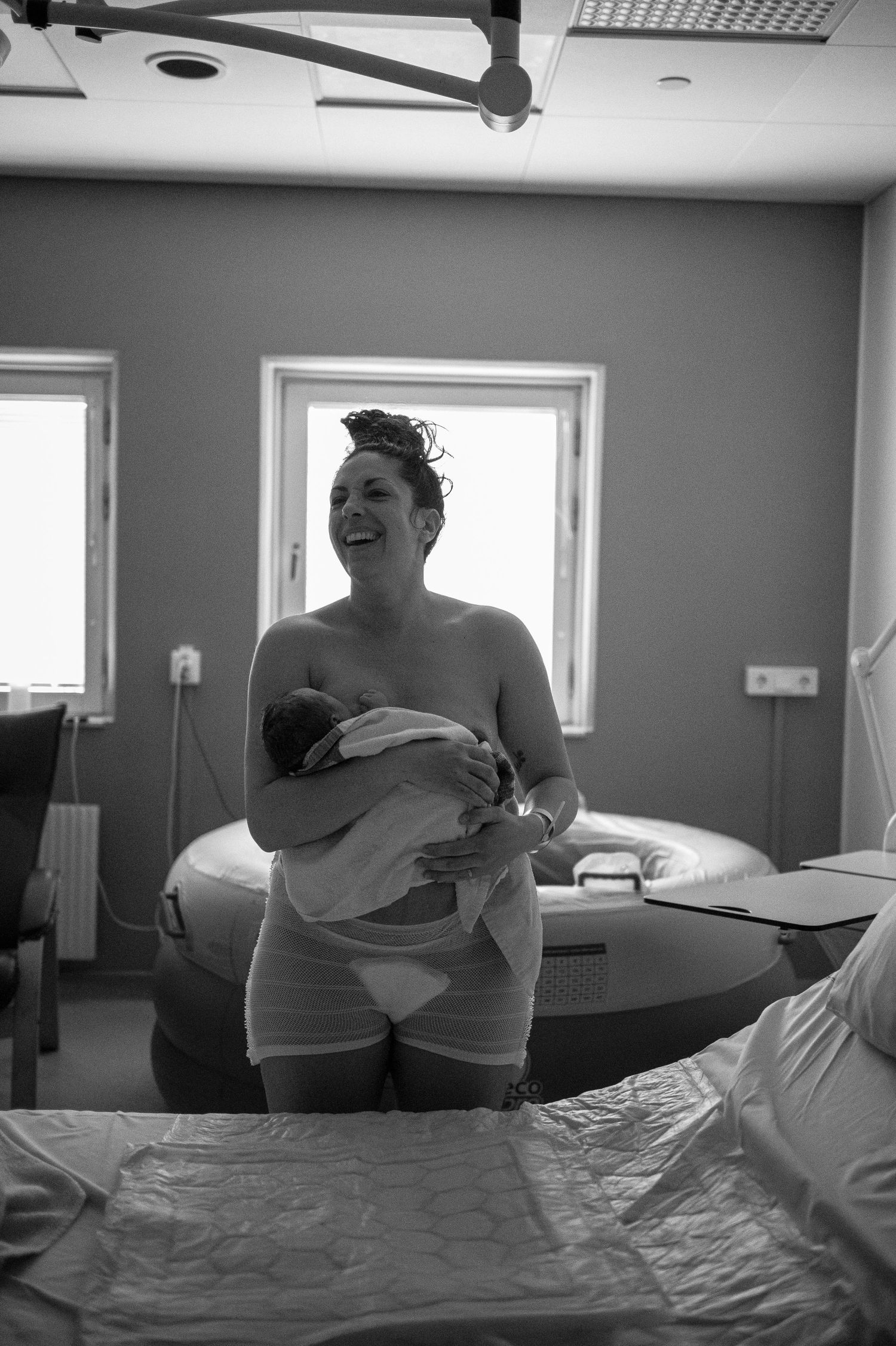 Black and white hospital room photo showing joyful new parent cradling newborn baby while standing near windows.