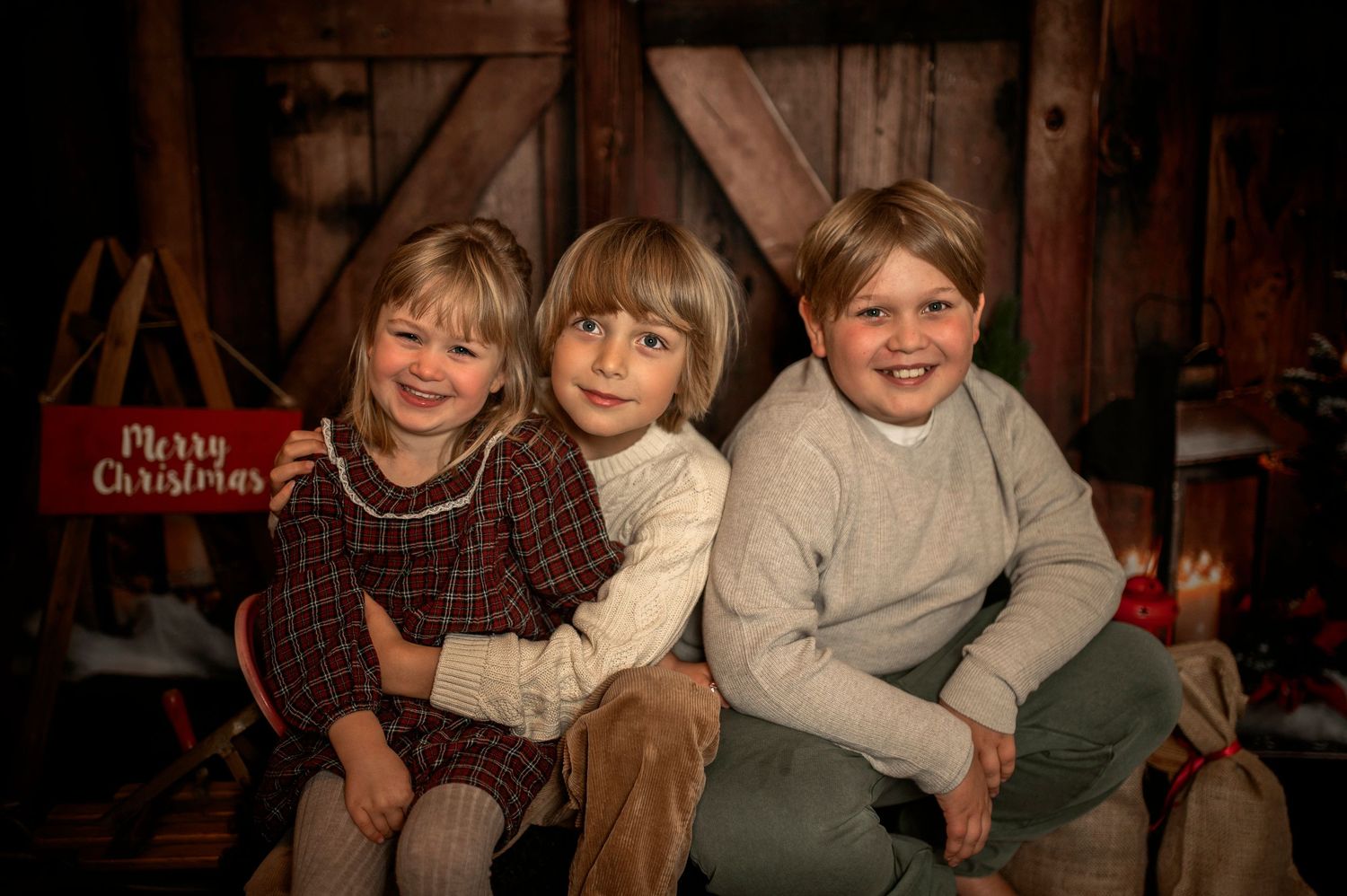 Three children pose together for a festive Christmas portrait in front of a rustic barn backdrop with holiday decorations.