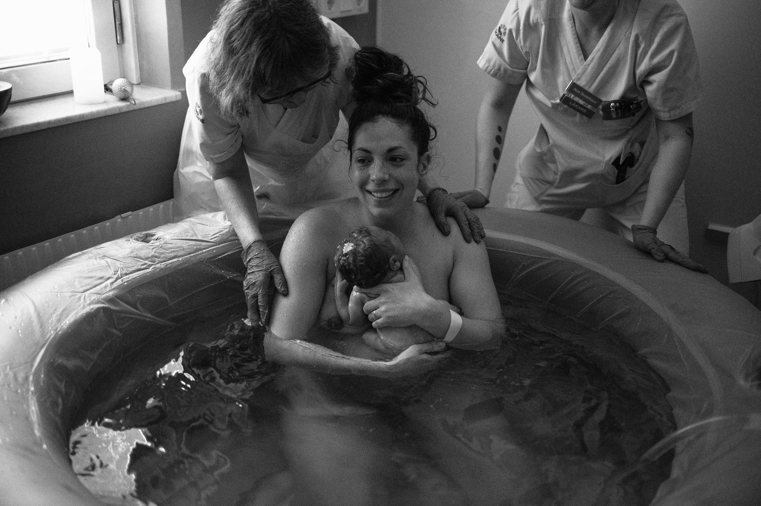 Black and white photo of woman in birthing pool holding newborn baby during water birth with midwives.