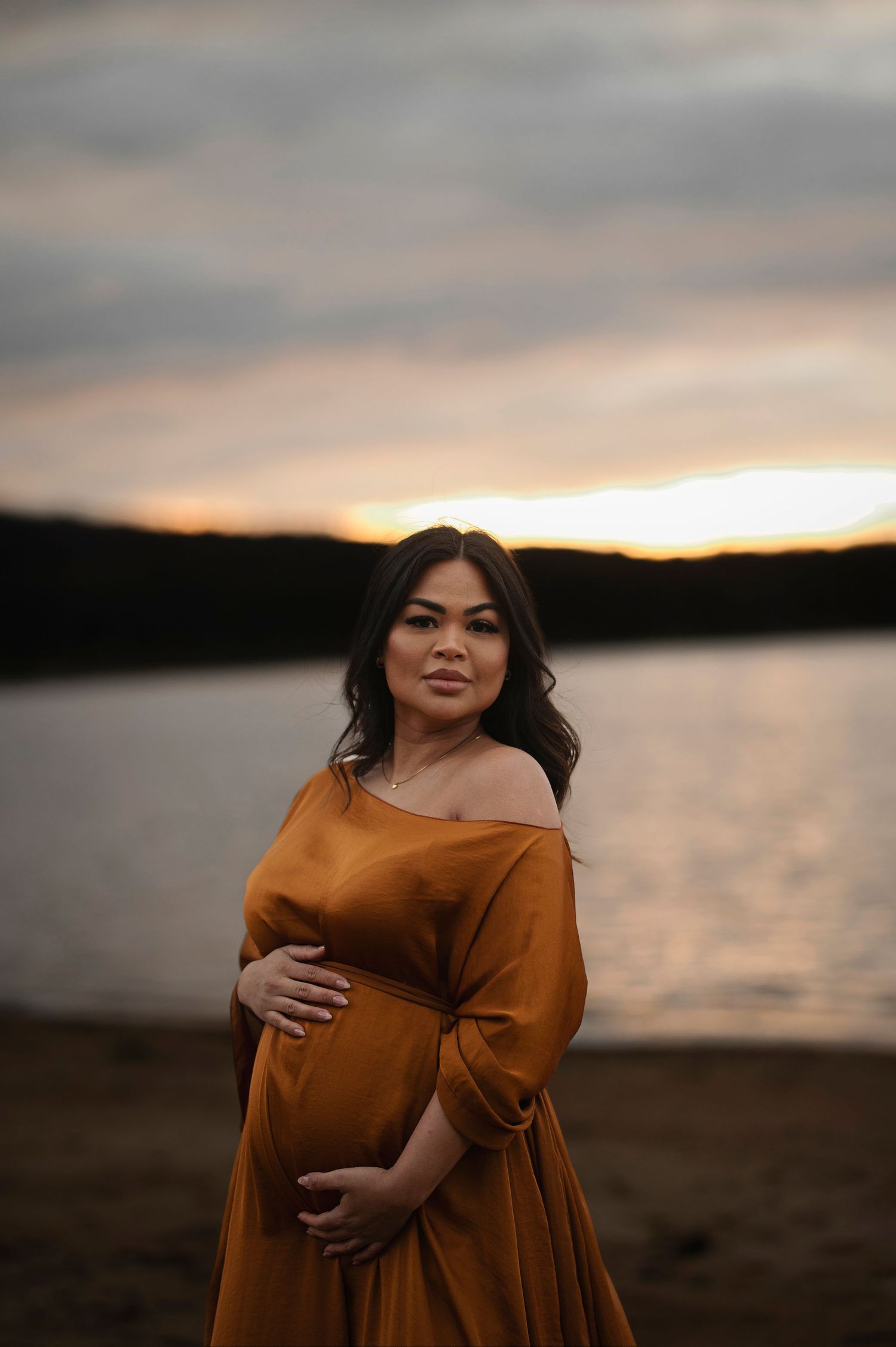 A pregnant woman in an off-shoulder orange dress poses by a lake during sunset.