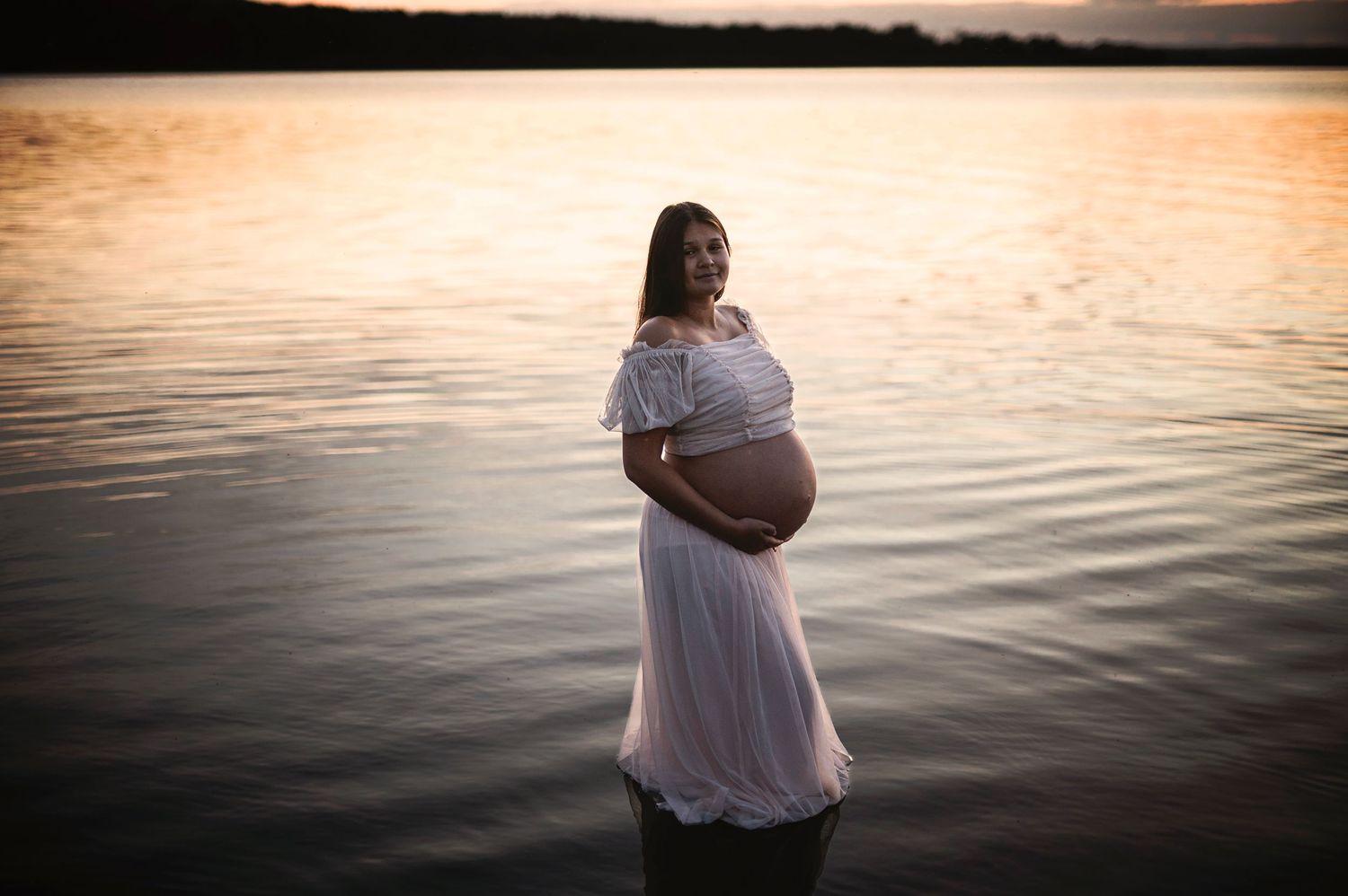 A serene maternity portrait at the water's edge during golden hour sunset.