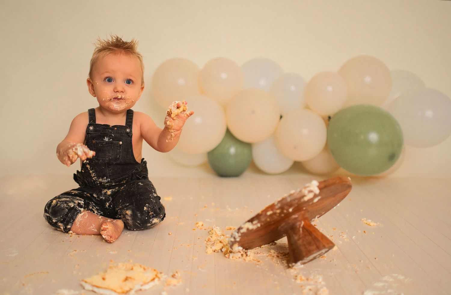 A festive cake smash scene with decorative balloons and scattered cake crumbs on the floor.