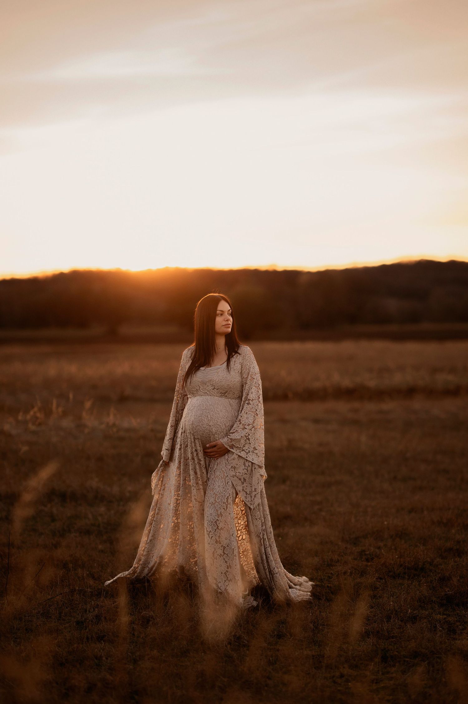 Gravidfotografering i Skåne med gravid kvinna i spetsklänning i gyllene fält vid solnedgången.