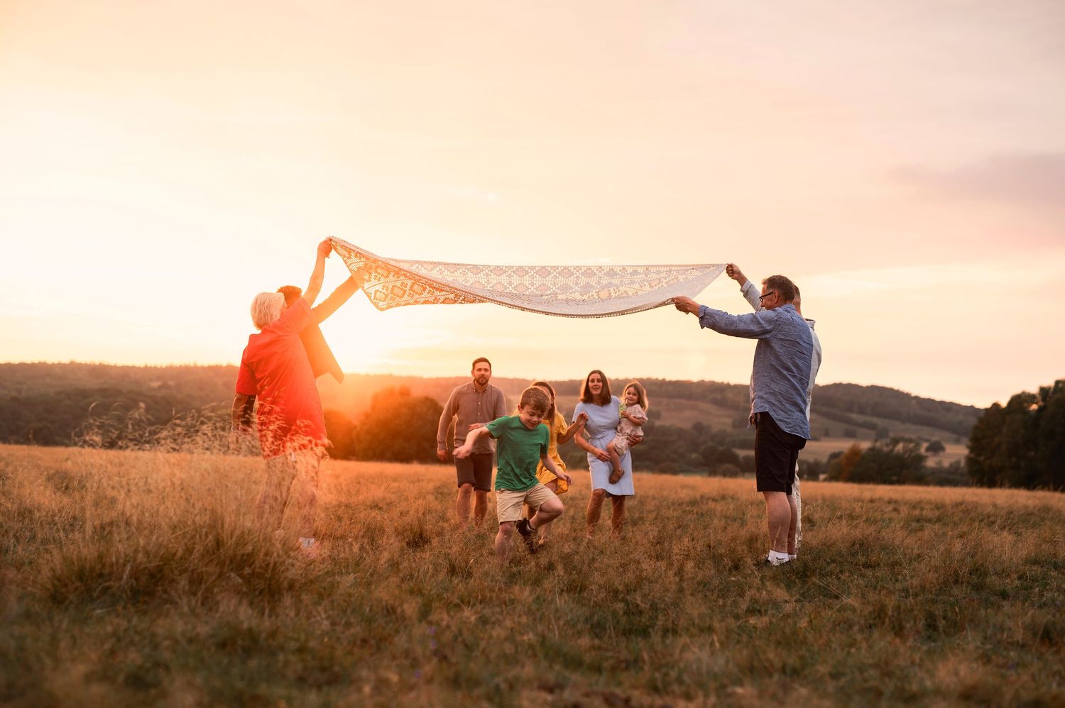 Familjefotografering i Skåne med flera generationer som leker med tyg i gyllene solnedgång på fält.