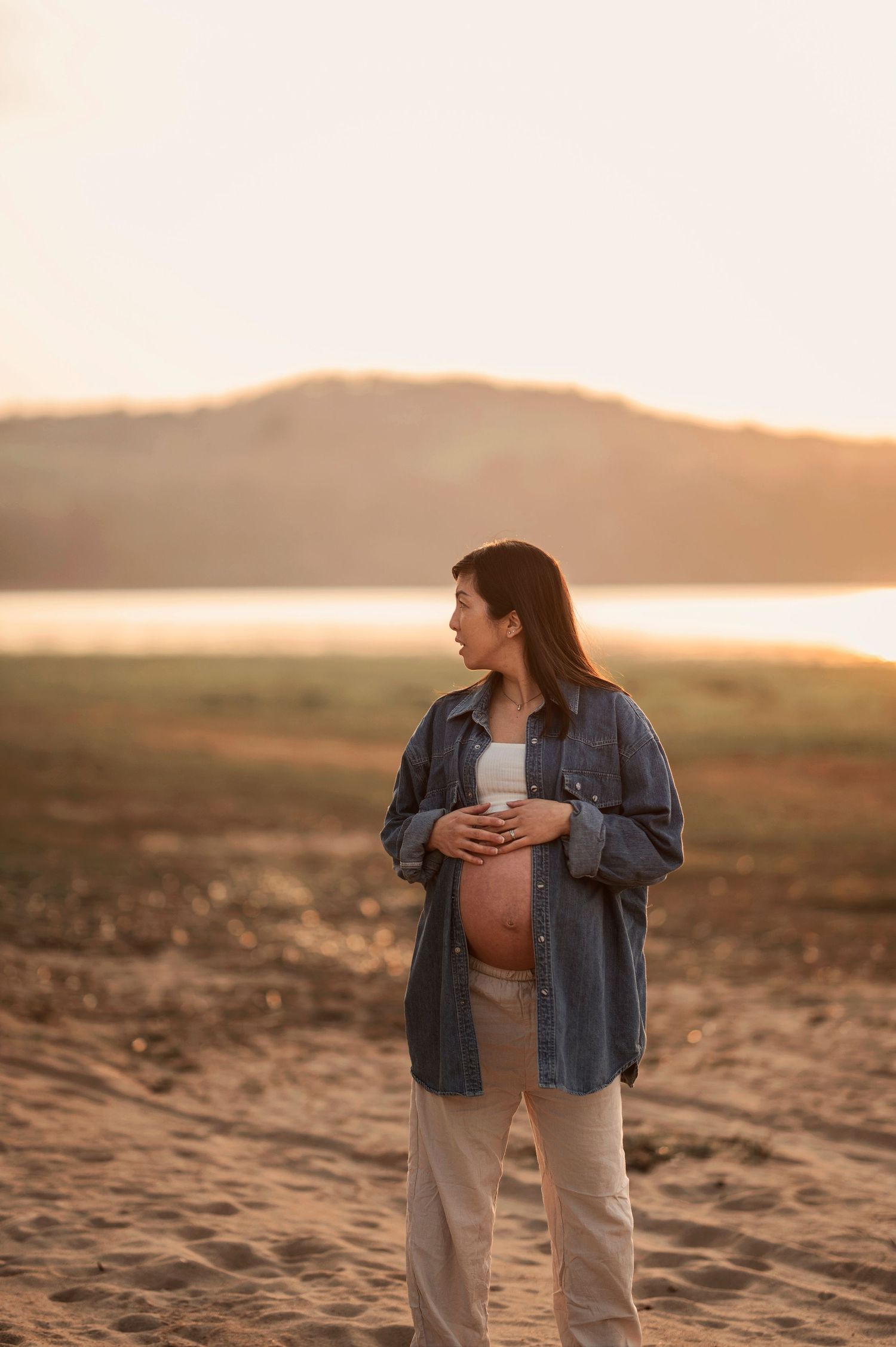 Gravid kvinna i jeansjacka fotograferad i ett öppet landskap under en gravidfotografering i Skåne i kvällsljus
