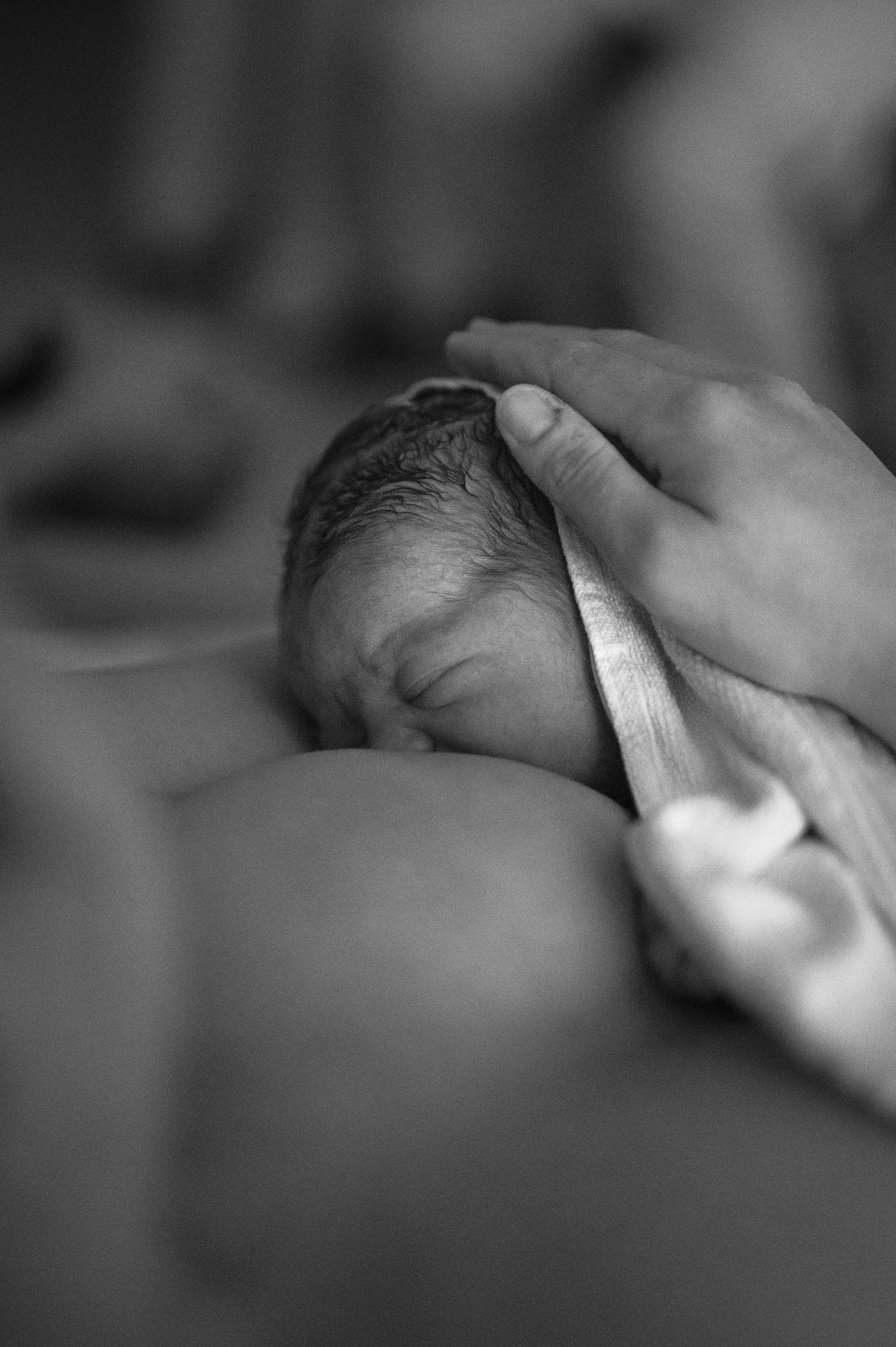A gentle hand touches a newborn baby's head while swaddled in soft cloth, captured in black and white.