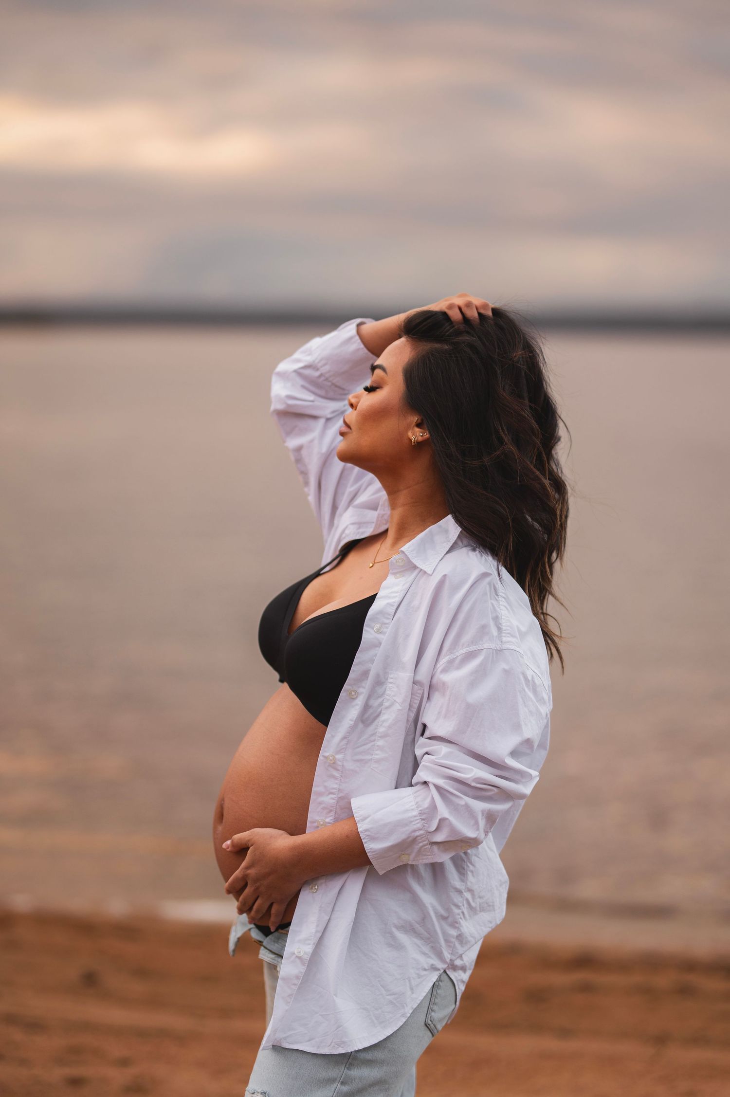 Expectant mother in white shirt and black top stands reflectively by a serene lake at sunset.