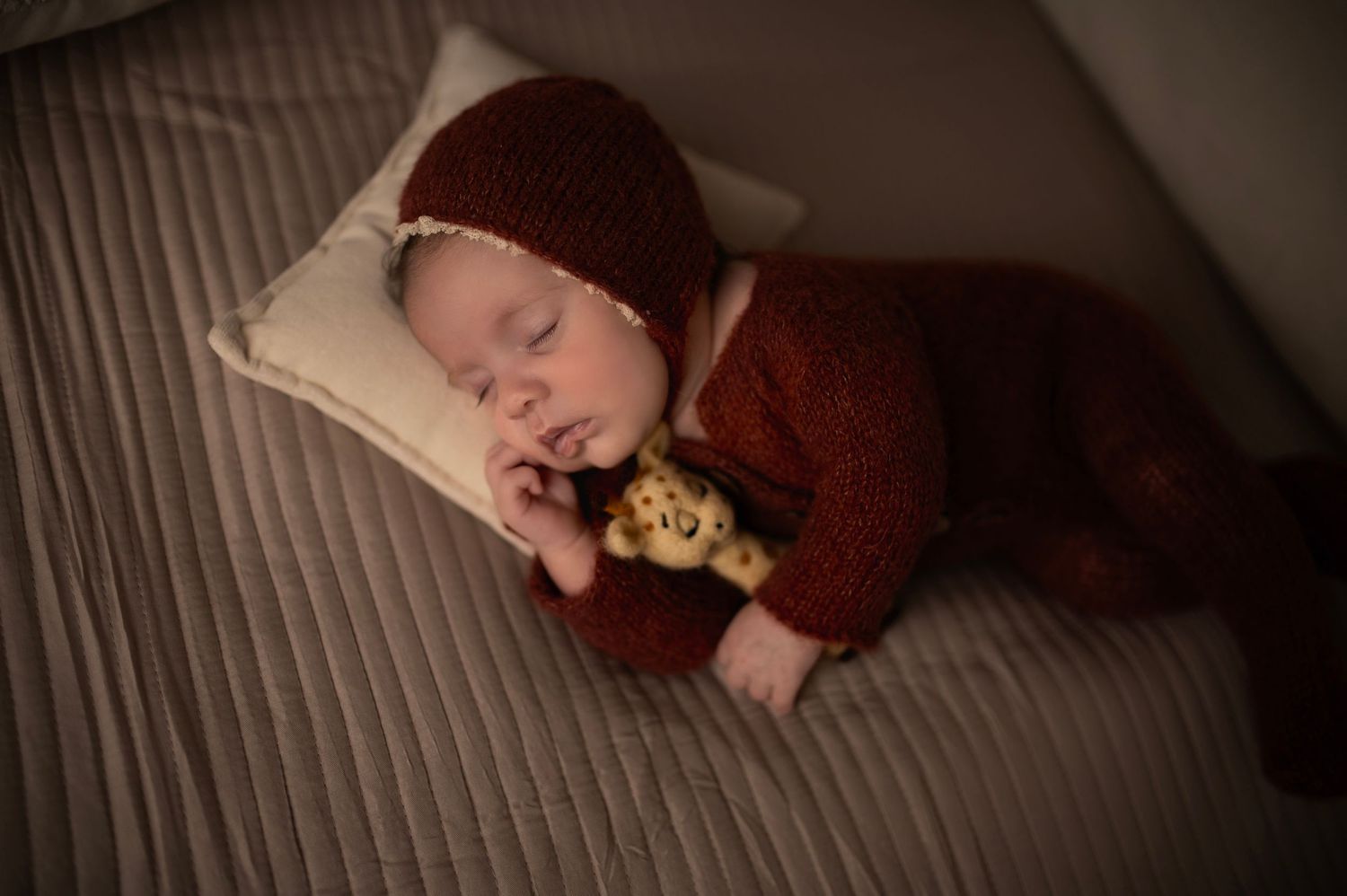 Peaceful infant in burgundy knit outfit sleeping on light cushion with small plush toy.