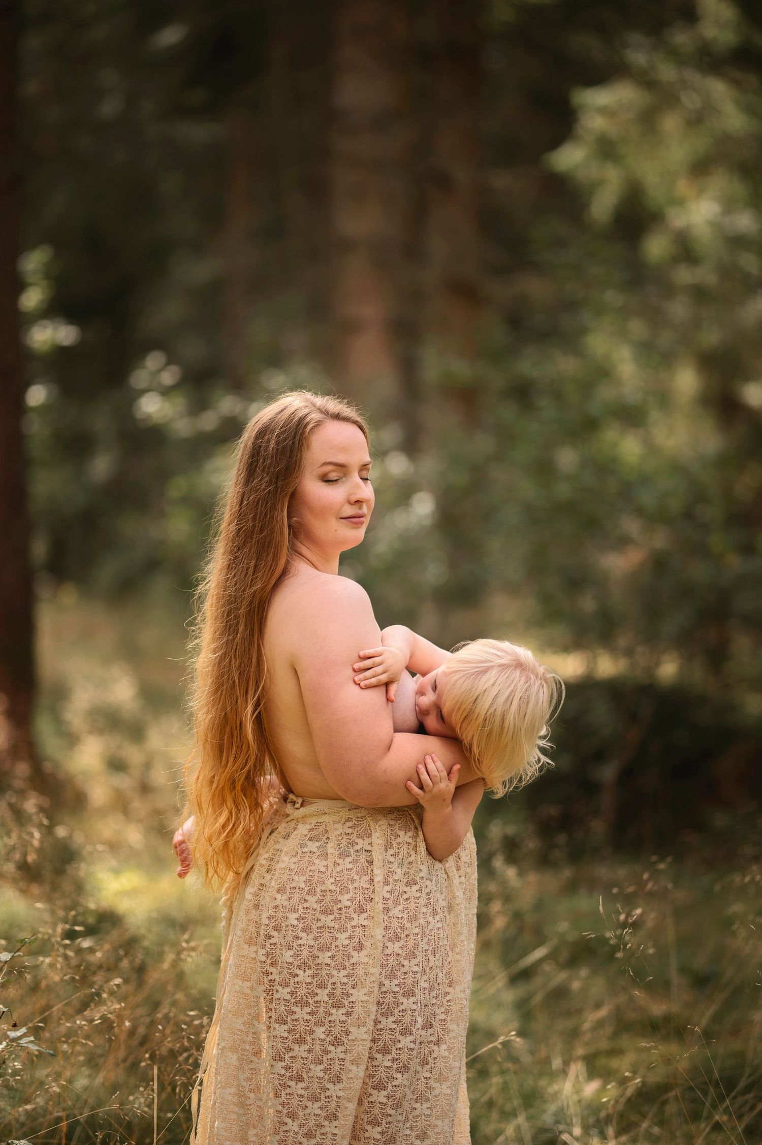 Moderskapsfotografering i Skåne med förälder som håller ett litet barn i spetsklänning i drömlik skogsmiljö.