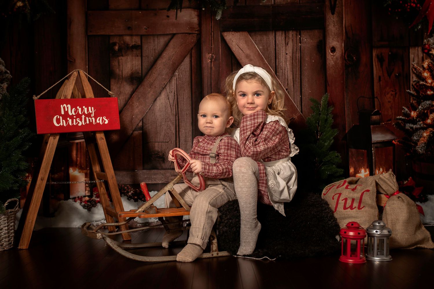 Two children pose on a sled near a Merry Christmas sign in a festive rustic holiday photo setting.