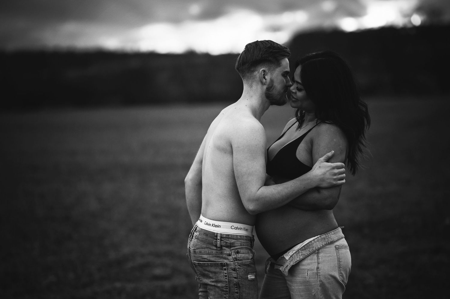 A dramatic black and white portrait of an expecting couple embracing outdoors.