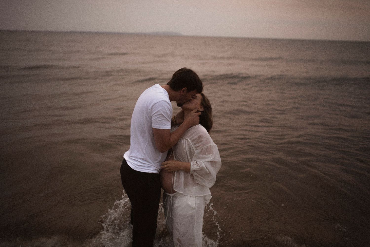 Couple shares intimate embrace while standing in ocean water during sunset.
