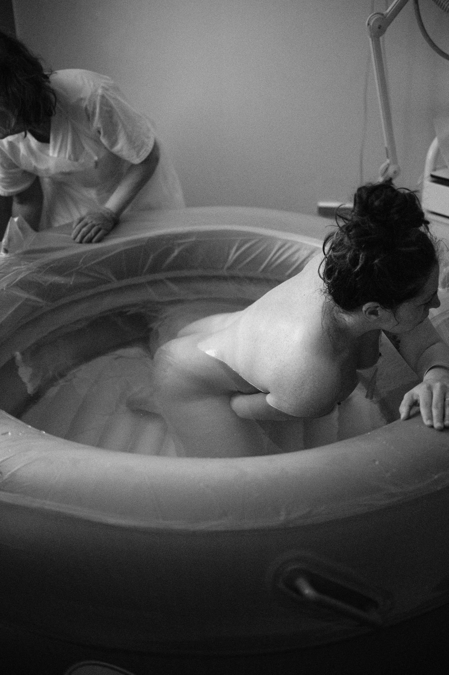 A person laboring in a birth pool during home water birth captured in moody black and white photography.