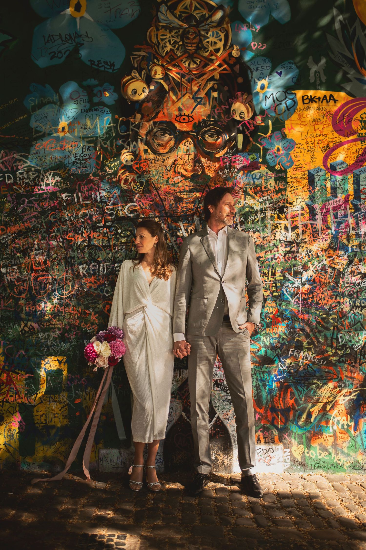 Couple in white dress and gray suit posing in front of colorful graffiti wall holding hands and pink bouquet. Elopement in Prauge